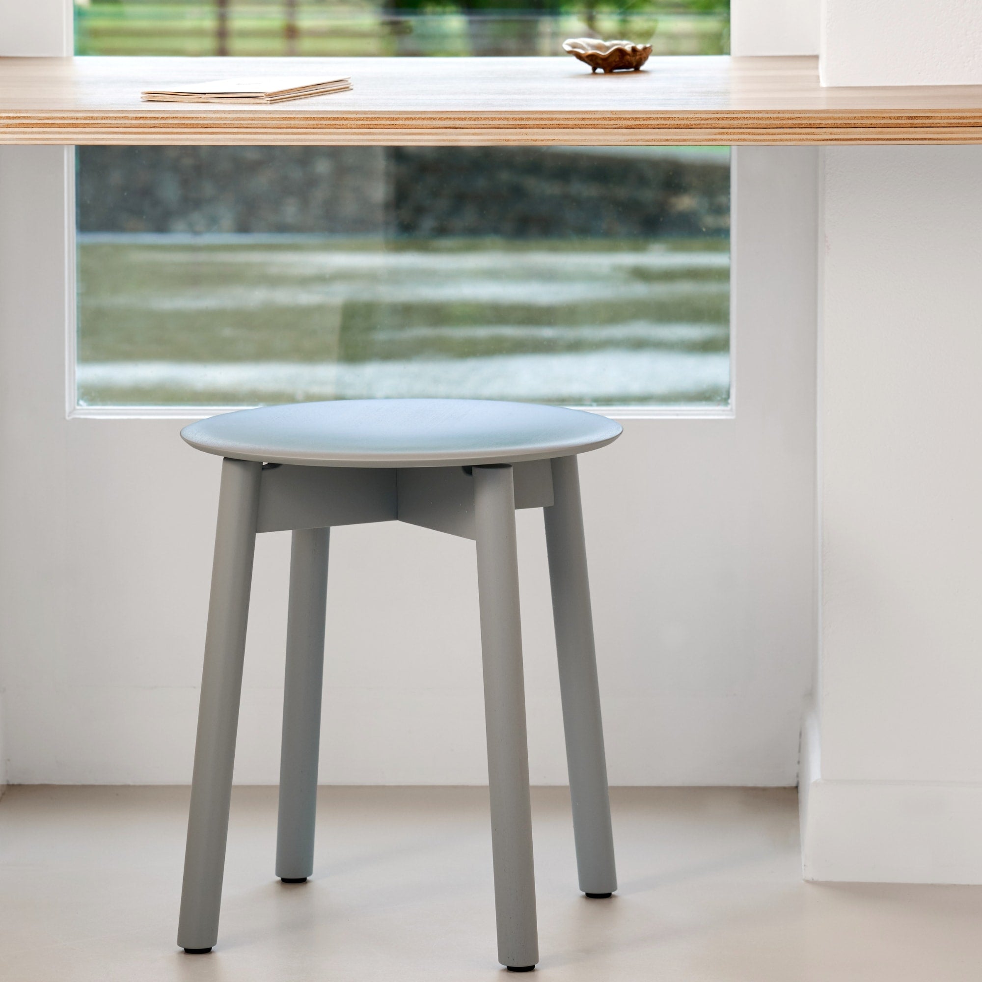 Tabouret Tiptoe en bois peint bleu clair, placé devant une baie vitrée et un bureau en contreplaqué
