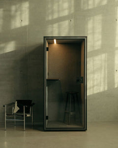 Cabine acoustique noire avec tabouret dans un espace minimaliste en béton, lumière rasante et fauteuil en cuir à gauche.