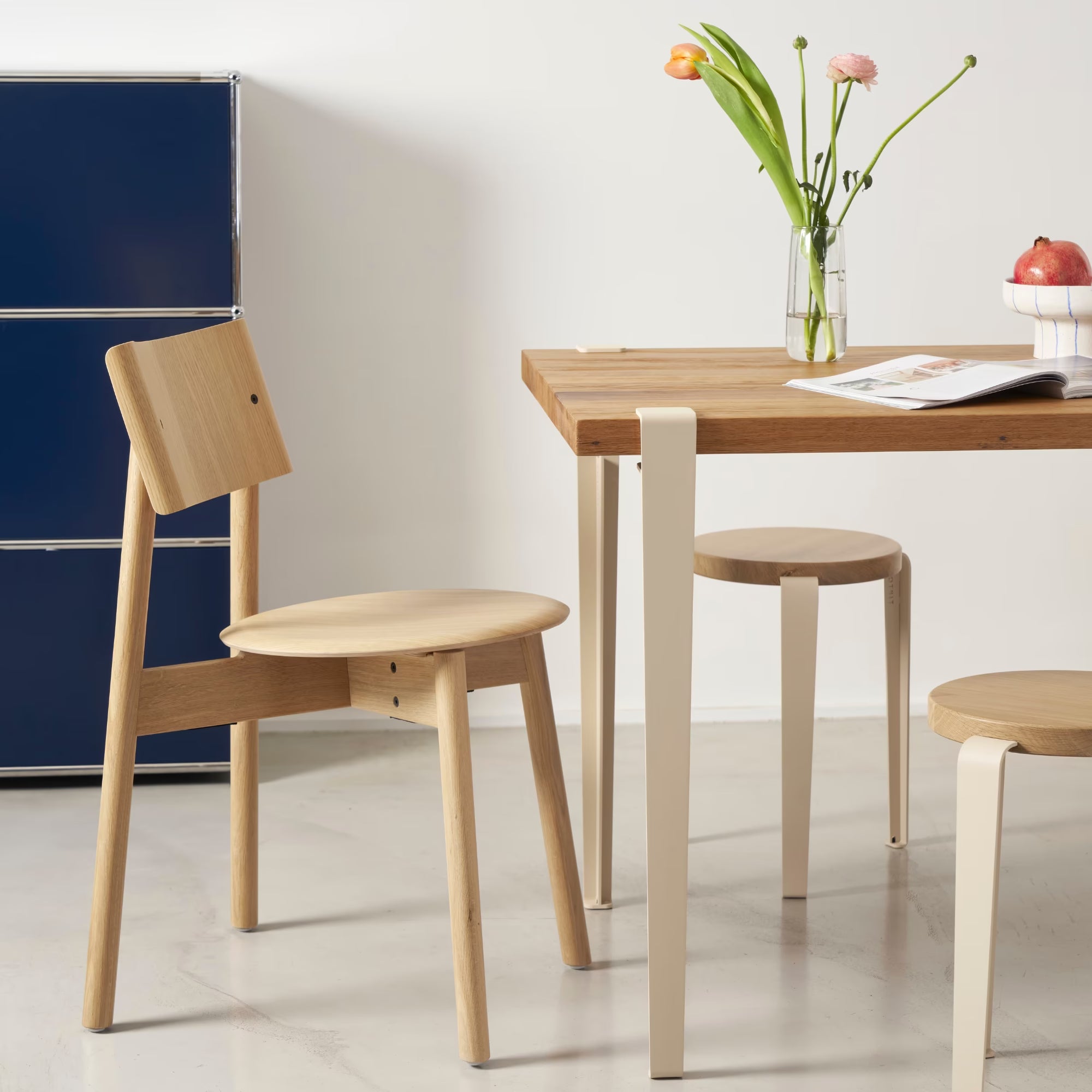 Scène repas minimaliste avec table en bois clair et pied métallique beige crème; chaise et deux tabourets en bois autour.