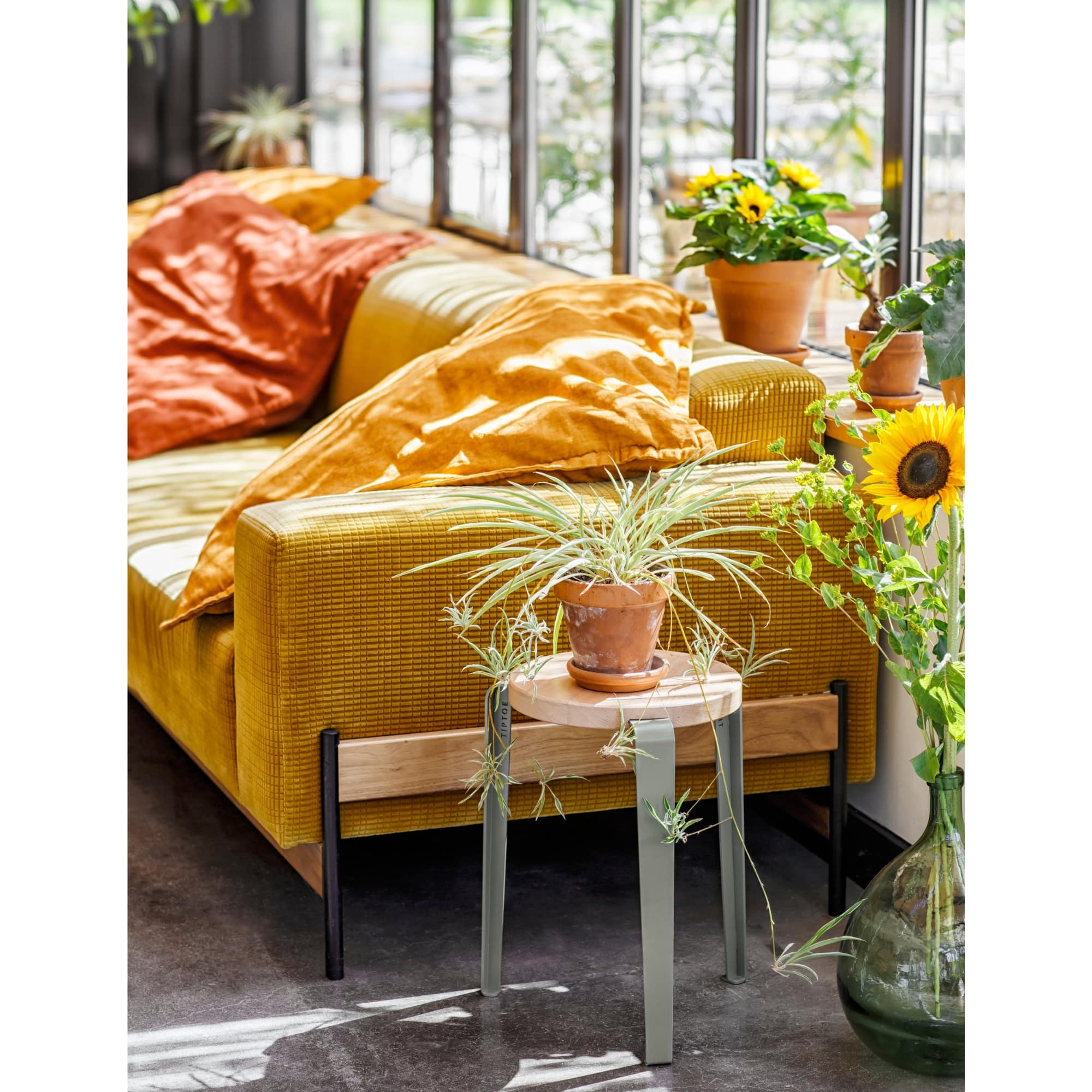 Tabouret Lou en chêne massif et acier gris clair, utilisé comme table d’appoint dans un salon baigné de lumière, avec plantes et canapé moutarde.