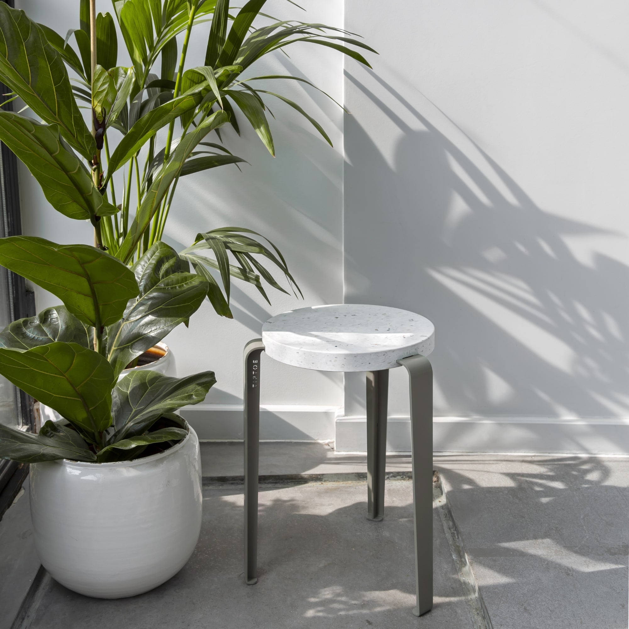 Tabouret Lou en plastique recyclé blanc et acier gris, placé près d’une plante en pot dans une pièce lumineuse