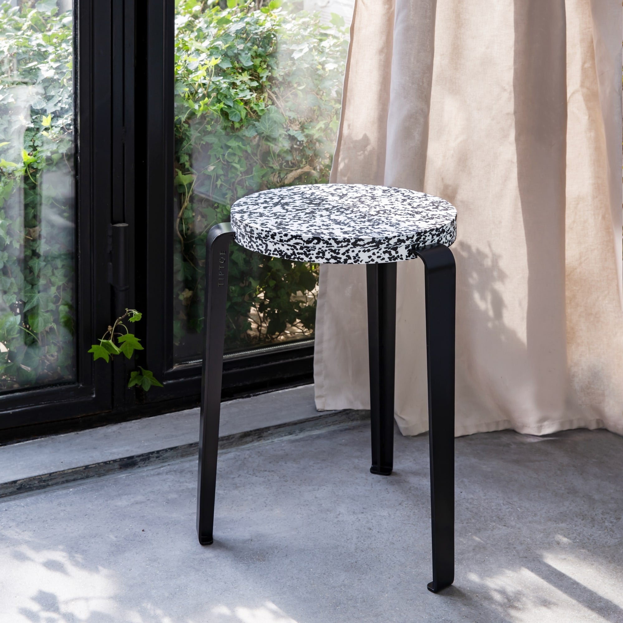 Tabouret Lou avec en plastique recyclé noir et blanc et pieds en acier noir, placé devant une baie vitrée et un rideau