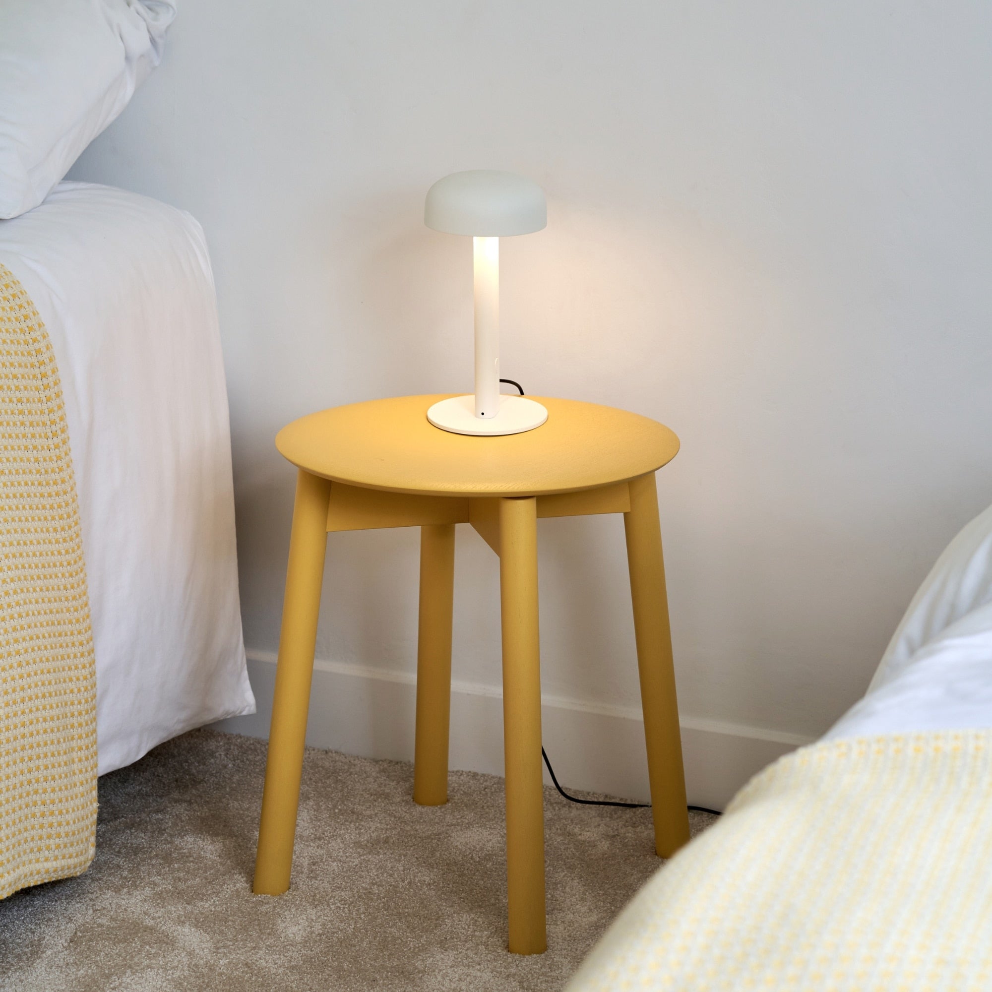 Tabouret Tiptoe en bois peint jaune utilisé comme table de chevet avec lampe blanche