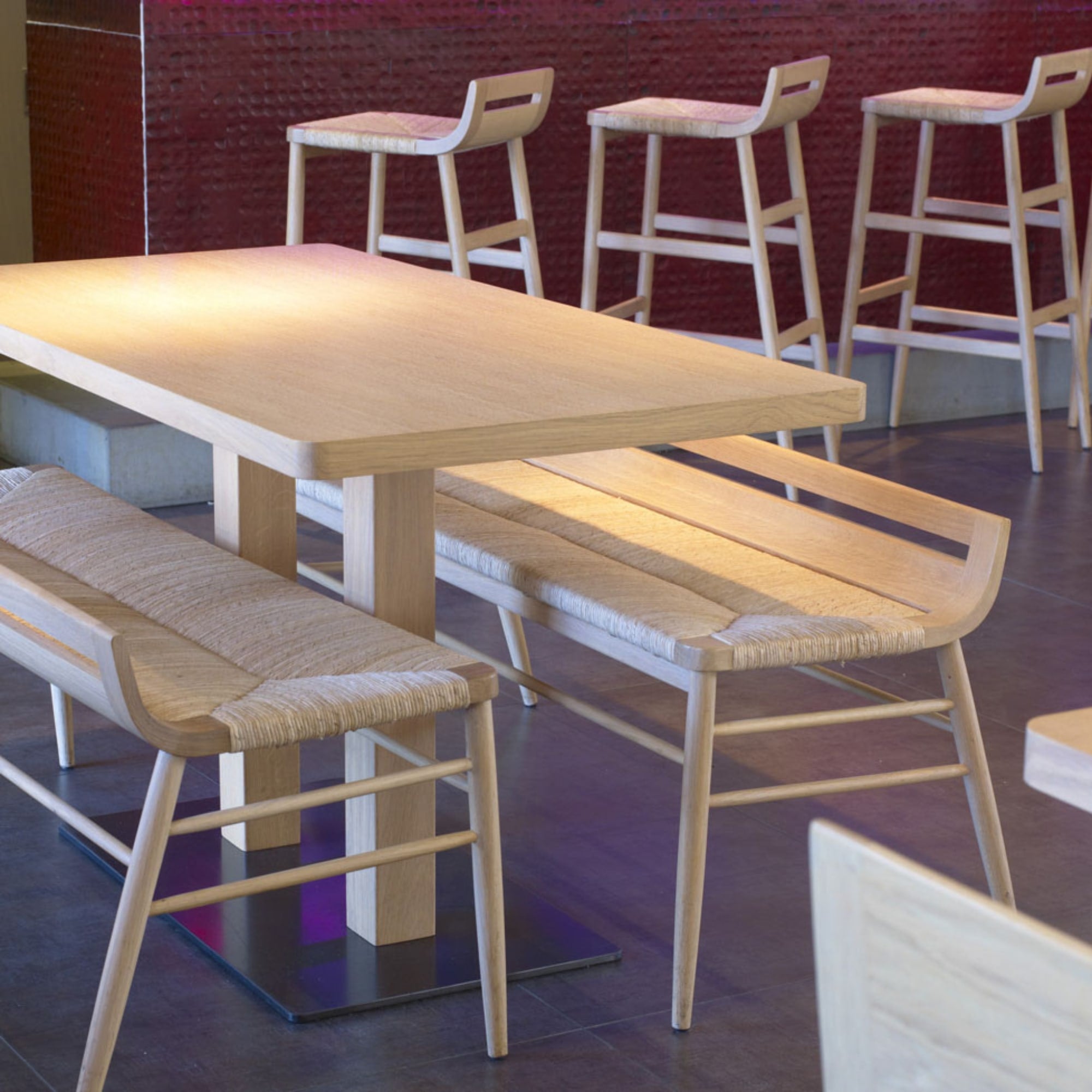 Bancs Kimua en bois et paille signés Alki, disposés de part et d’autre d’une table en chêne clair dans un restaurant