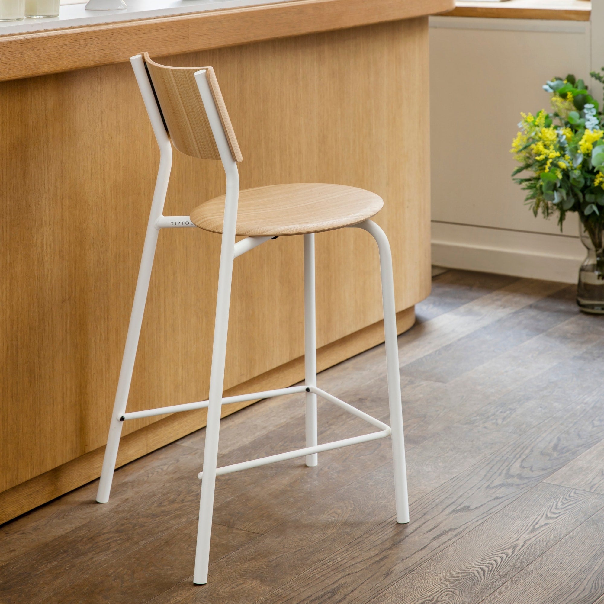 Chaise mi-hauteur avec structure en métal blanc et assise en bois clair devant un comptoir arrondi en bois dans un intérieur chaleureux avec bouquet de fleurs.