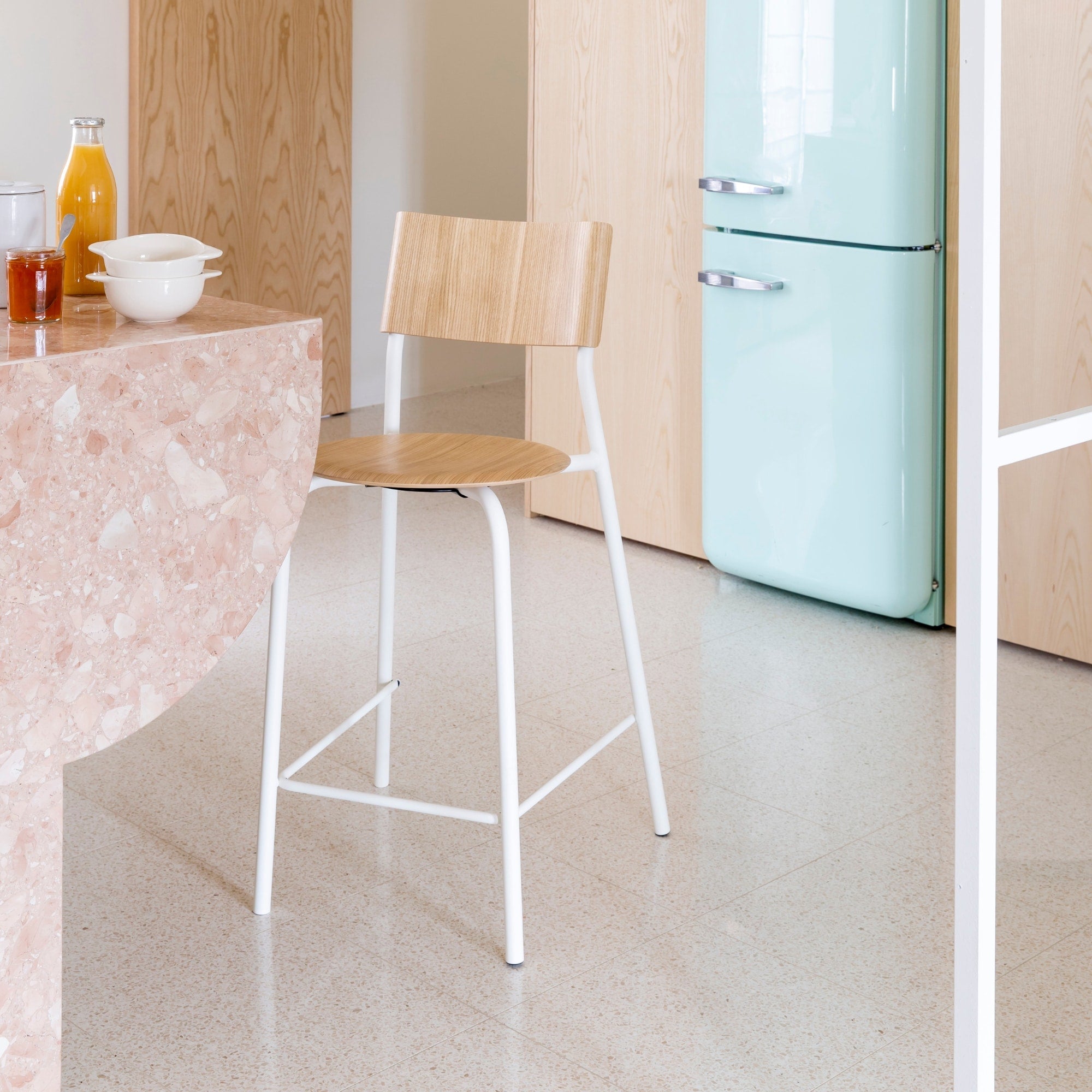 Chaise mi-hauteur blanche avec assise et dossier en bois clair devant un îlot de cuisine en marbre rose dans un intérieur lumineux avec réfrigérateur vert pastel en arrière-plan.
