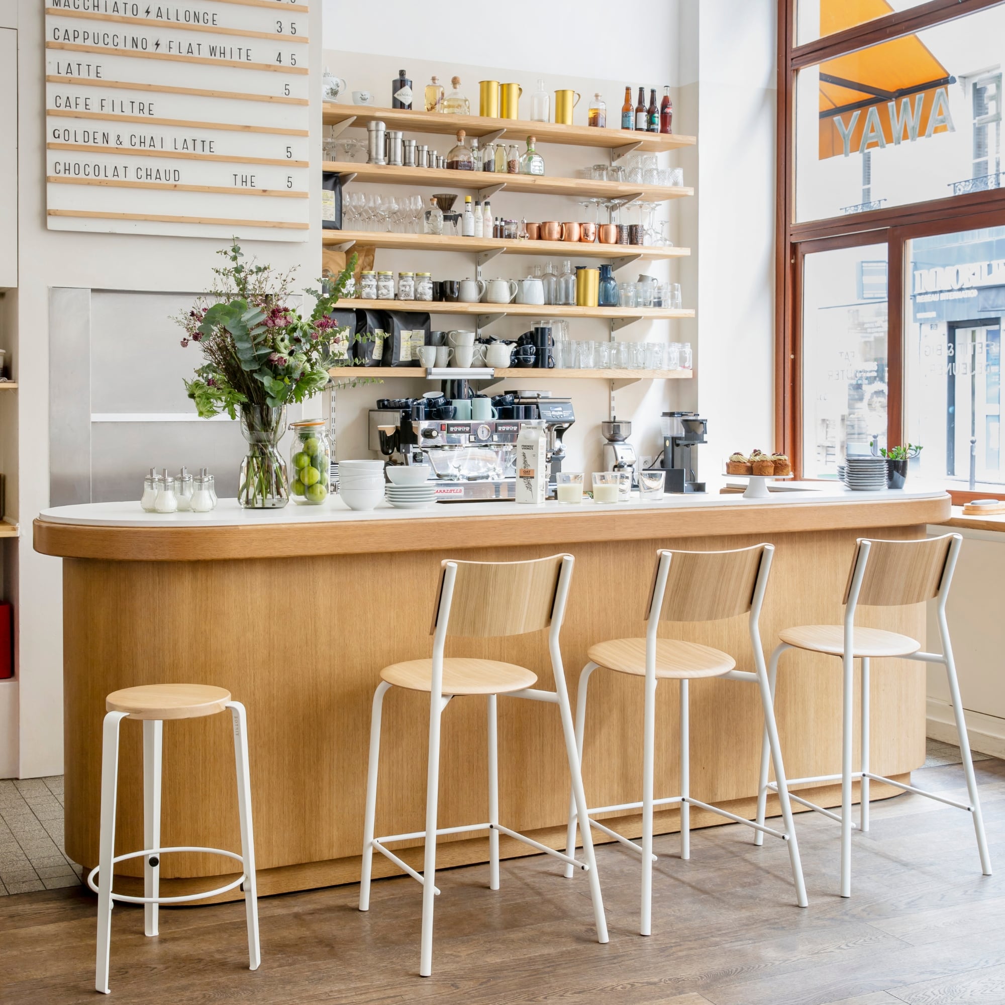 Ensemble de trois chaises mi-hauteur et un tabouret blans avec assise et dossier en bois clair alignées devant un comptoir de café en bois avec vaisselle et bouquets de fleurs sur les étagères en arrière-plan.