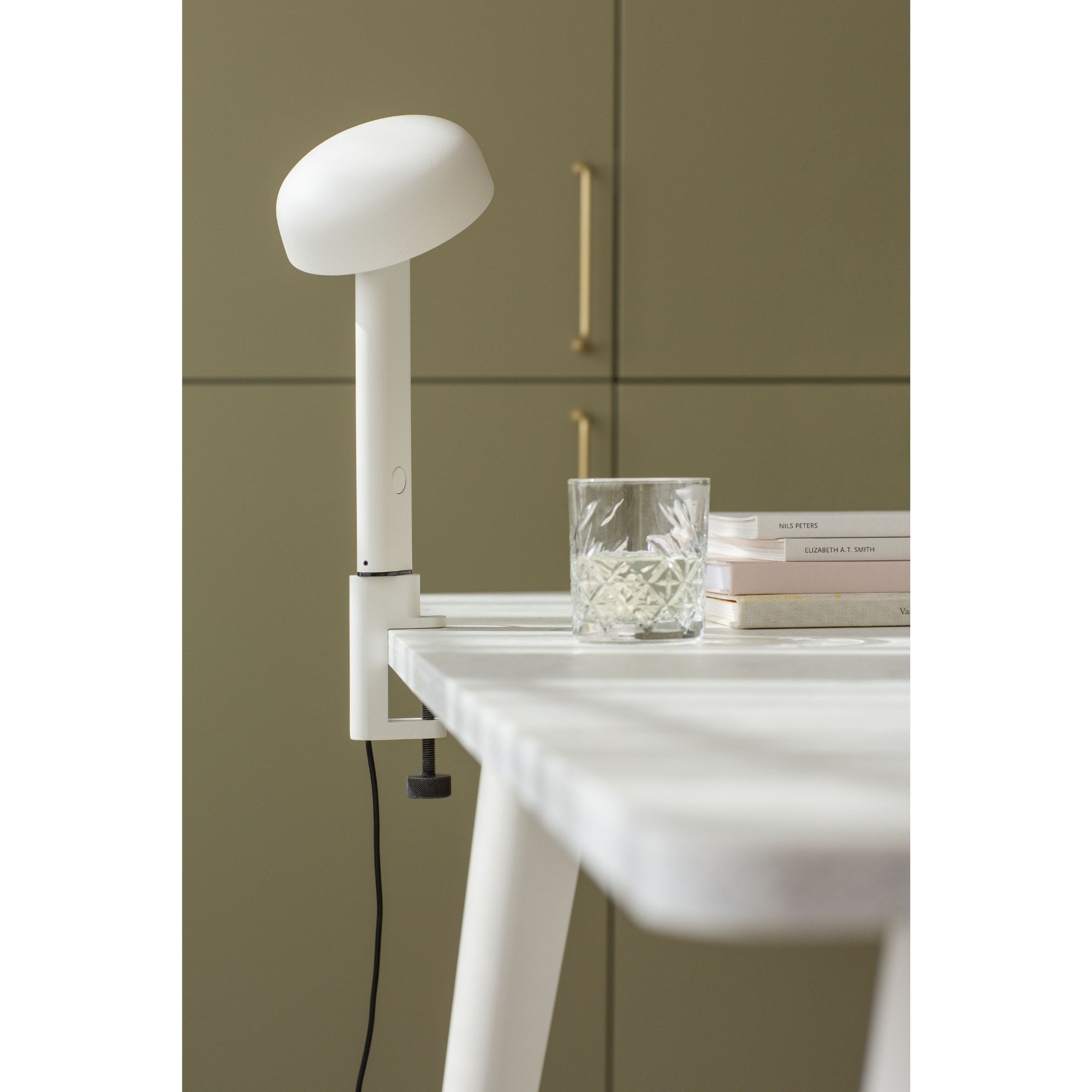 Lampe à pince blanche, modèle Nod de Tiptoe, vue latérale. Le bras fin soutient un abat-jour circulaire légèrement orienté vers le bas. Un verre et des livres sont visibles sur la table.