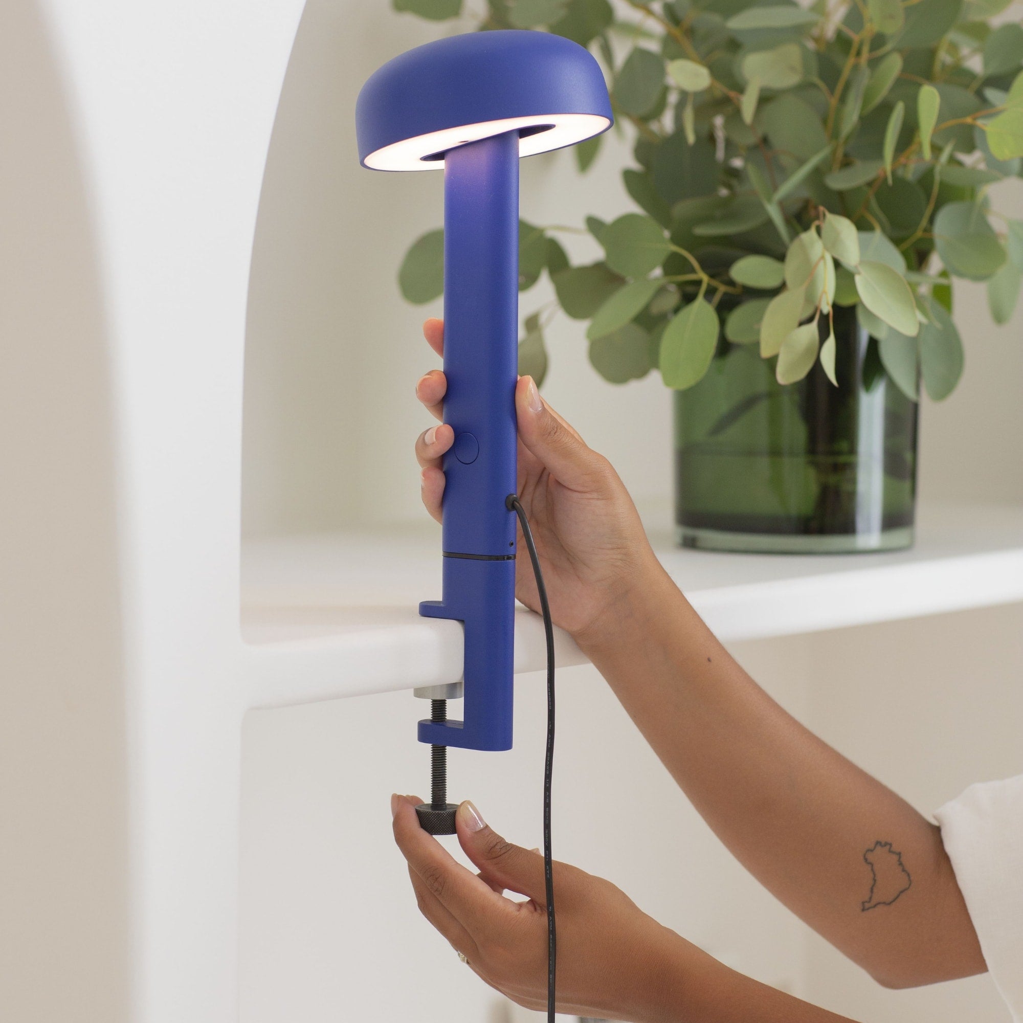Lampe à pince bleue, modèle Nod de Tiptoe, vue en gros plan montrant une main ajustant la lampe. Le bras est fixé sur une étagère, avec l’abat-jour circulaire allumé, orienté vers le bas, et la lumière visible.