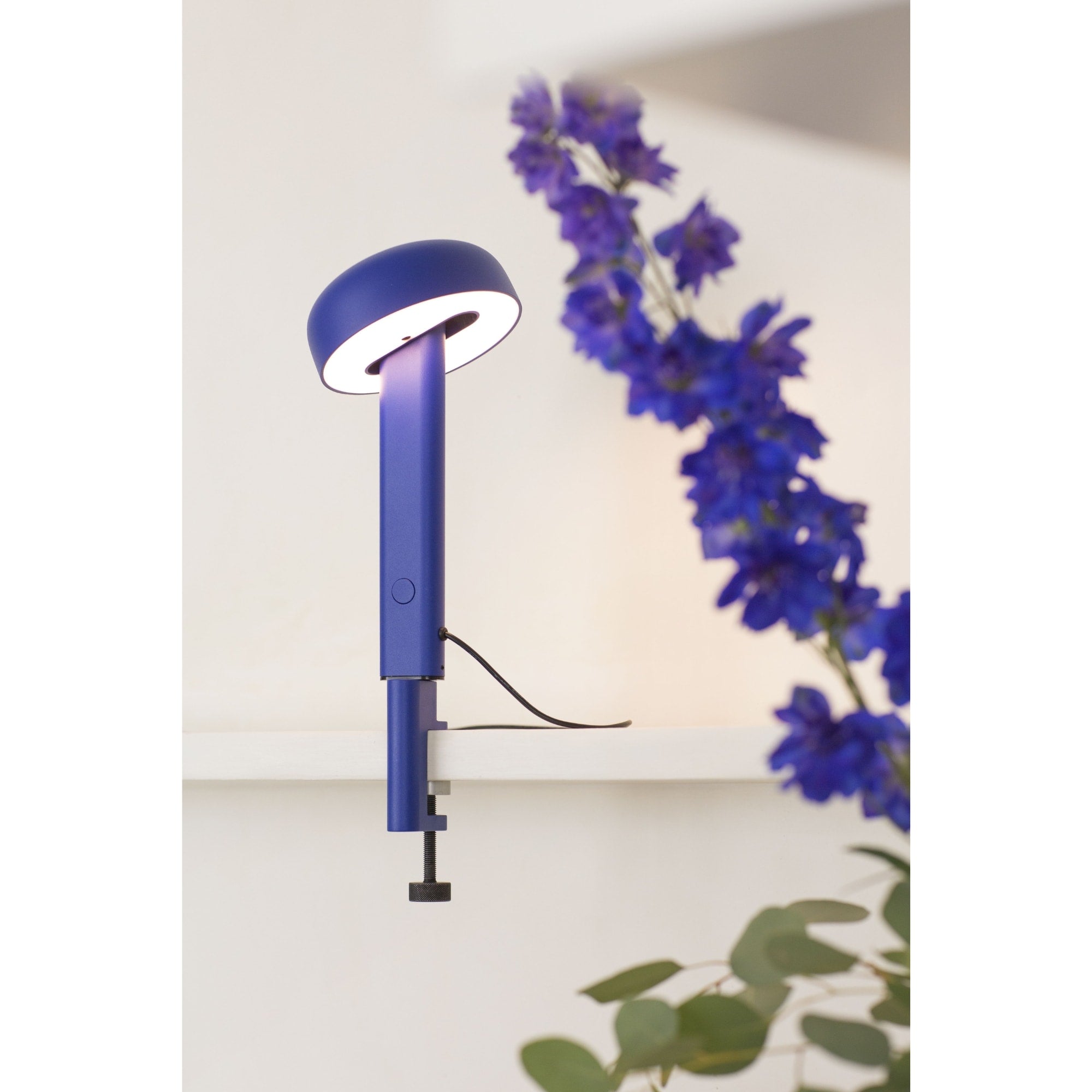 Lampe à pince bleue, modèle Nod de Tiptoe, allumée. Vue latérale avec un bras fin soutenant un abat-jour circulaire, la lumière émise éclaire une zone bien définie. Des fleurs bleues et des feuilles décorent l’arrière-plan.