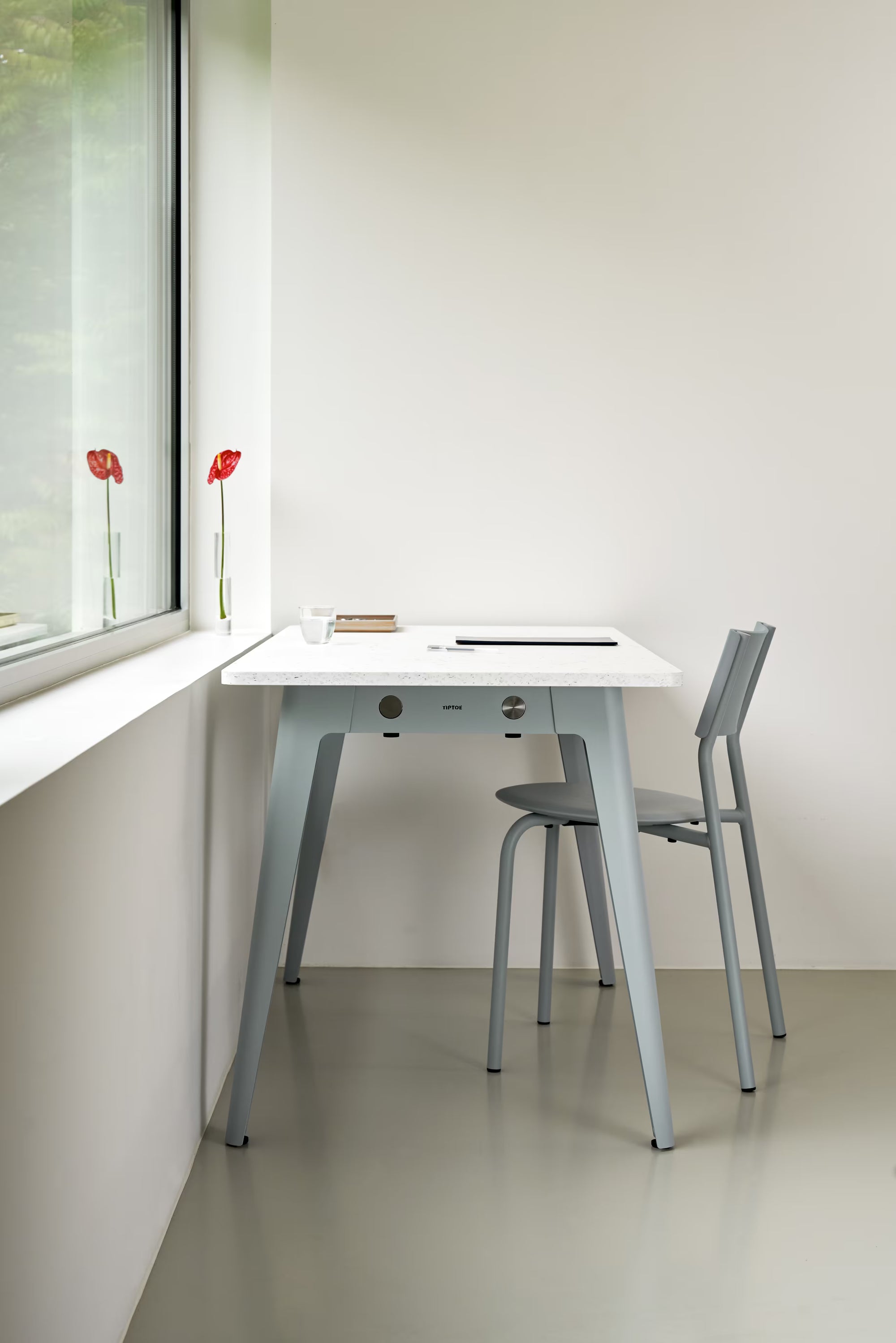 Vue d’un coin bureau près d’une grande fenêtre, table claire aux pieds gris, deux tabourets, mur blanc et coquelicots sur l’appui
