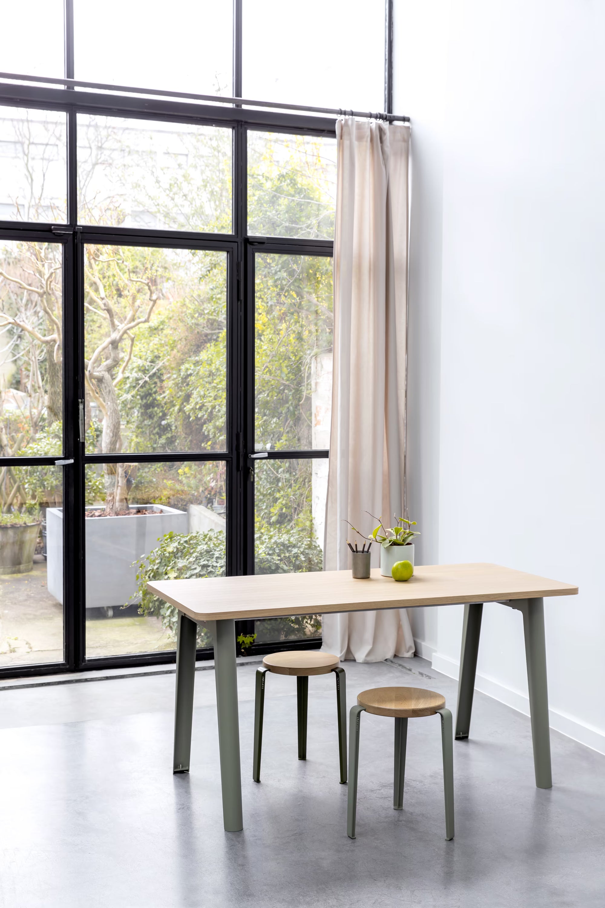 Pièce lumineuse avec grande verrière donnant sur un jardin, table chêne clair aux pieds vert gris et deux tabourets assortis.