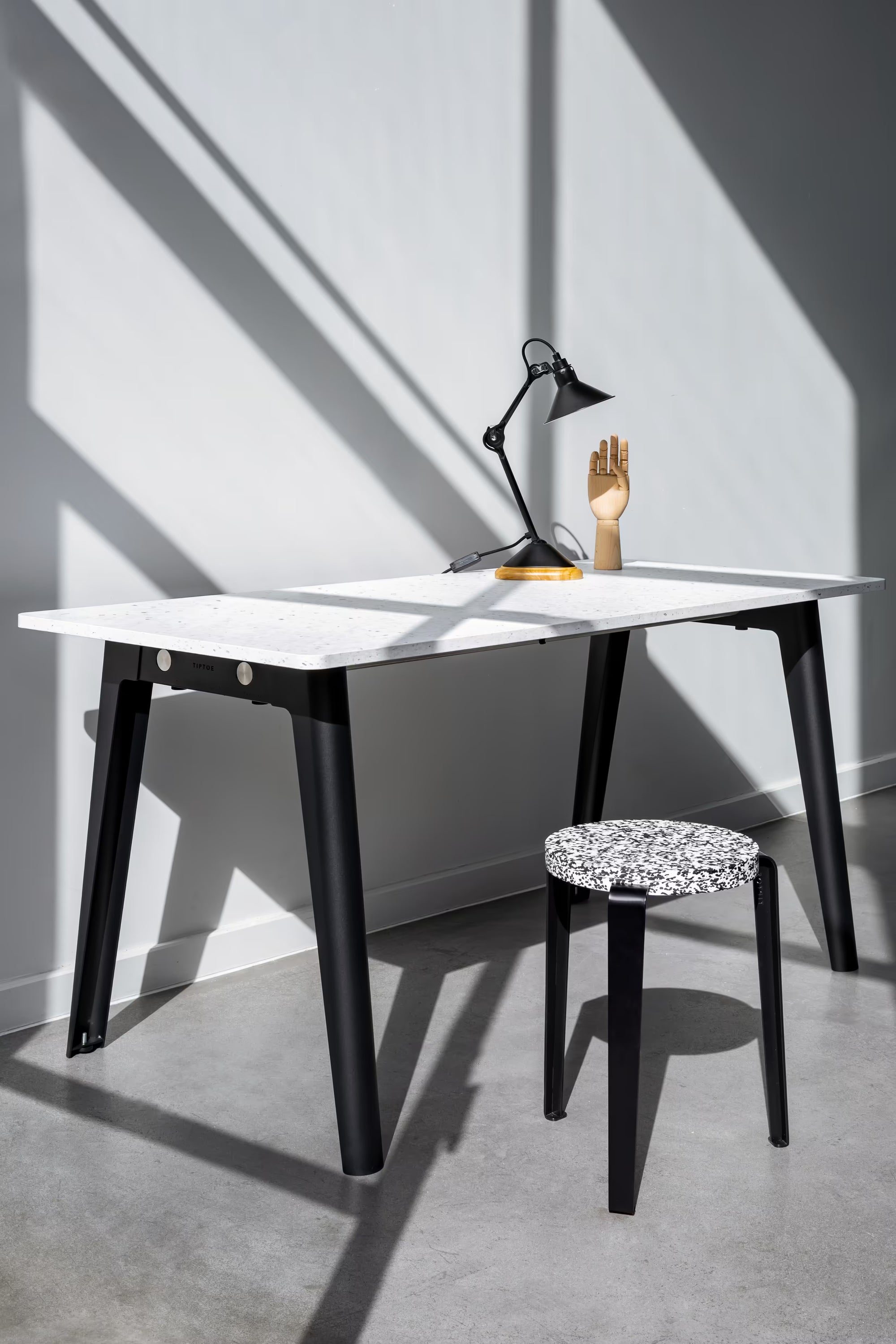 Bureau minimal, plateau blanc moucheté, pieds noirs, lampe posée et tabouret assorti, lumière rasante et ombres graphiques.