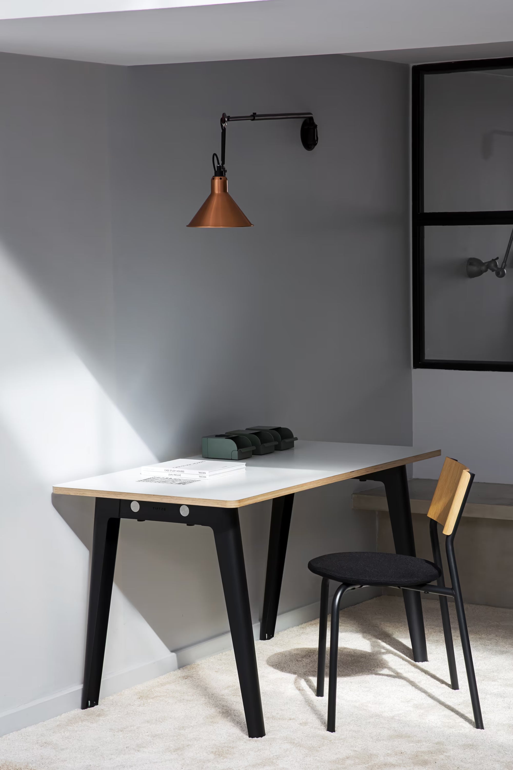 Bureau blanc aux pieds noirs avec chaise assortie, installé contre un mur gris avec applique cuivre et fenêtre intérieure.