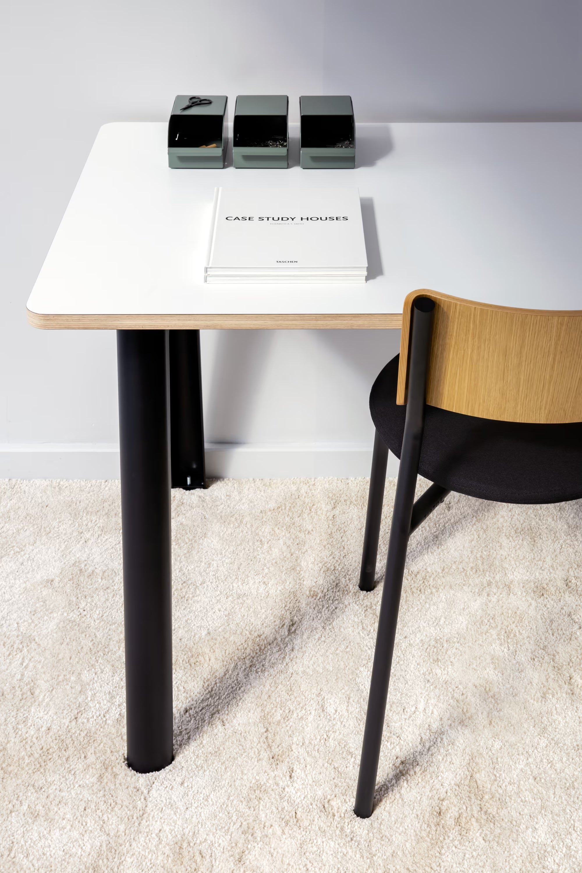 Vue rapprochée d’un bureau blanc avec pieds noirs et chaise bois, posé sur tapis clair avec objets et livre en surface.