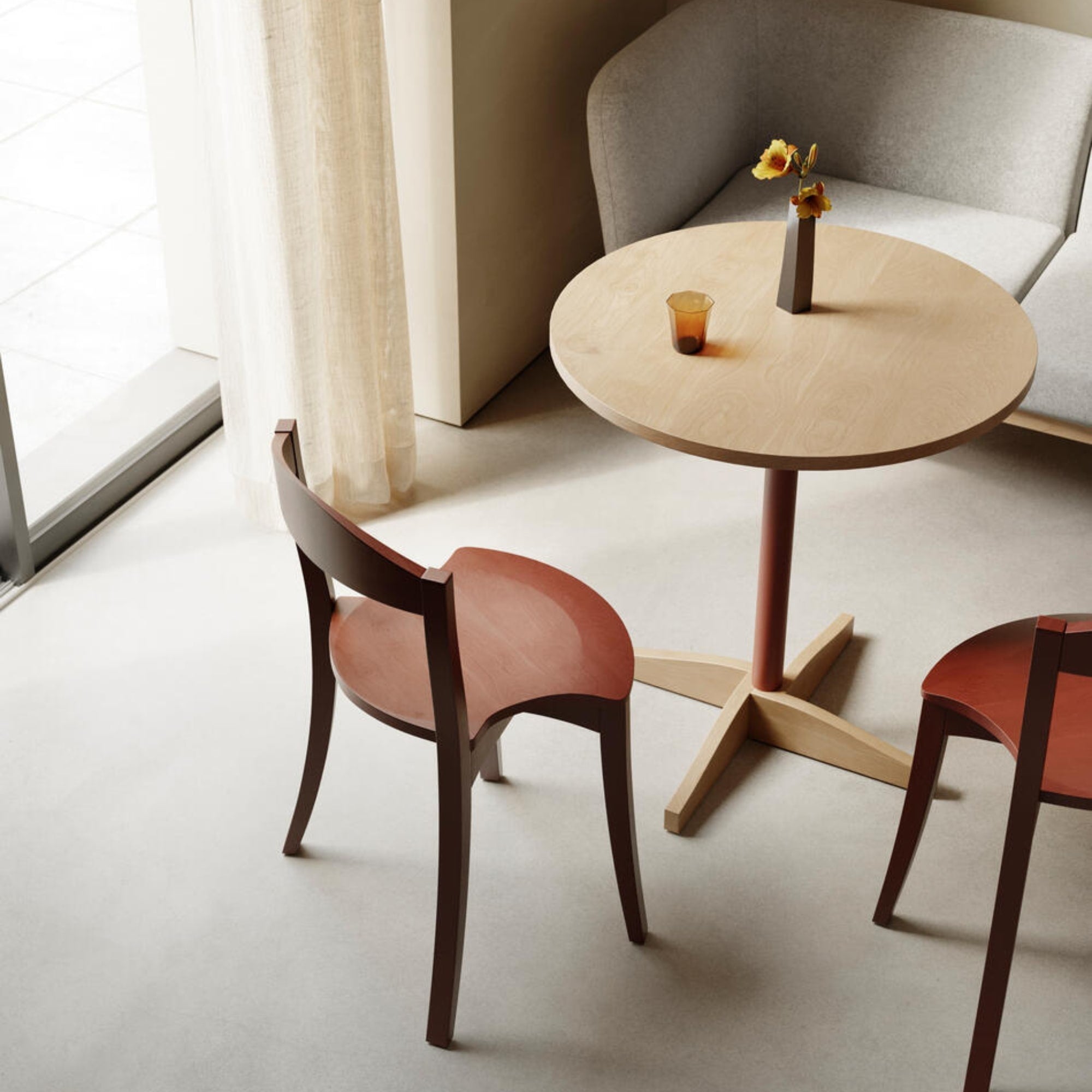 Deux chaises Bibolina rouges autour d’une table ronde claire, dans un coin salon lumineux et épuré.