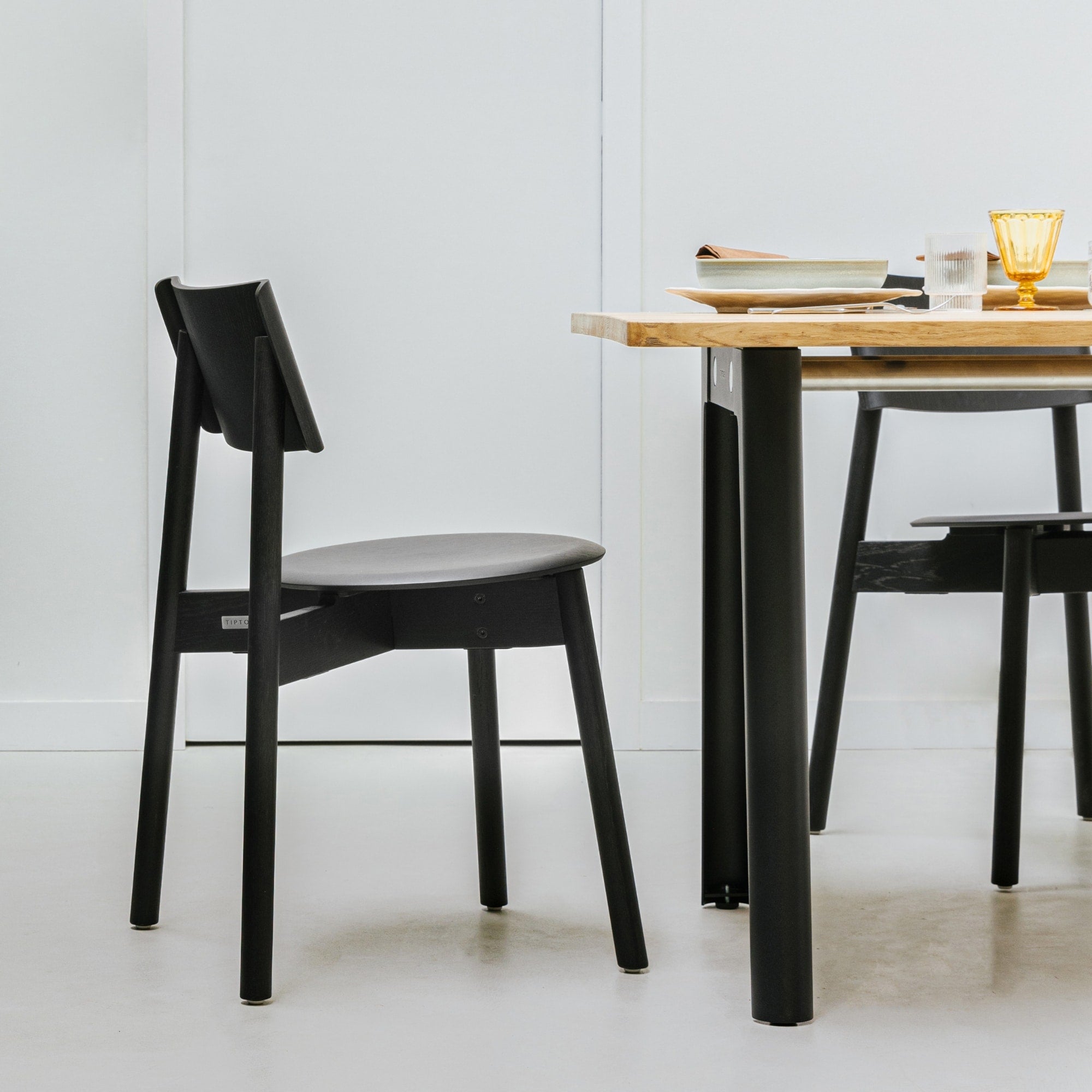 Chaise SSD Full Wood de Tiptoe en chêne teinté noir, placée à côté d’une table en bois clair aux pieds noirs, dans un décor sobre et lumineux.
