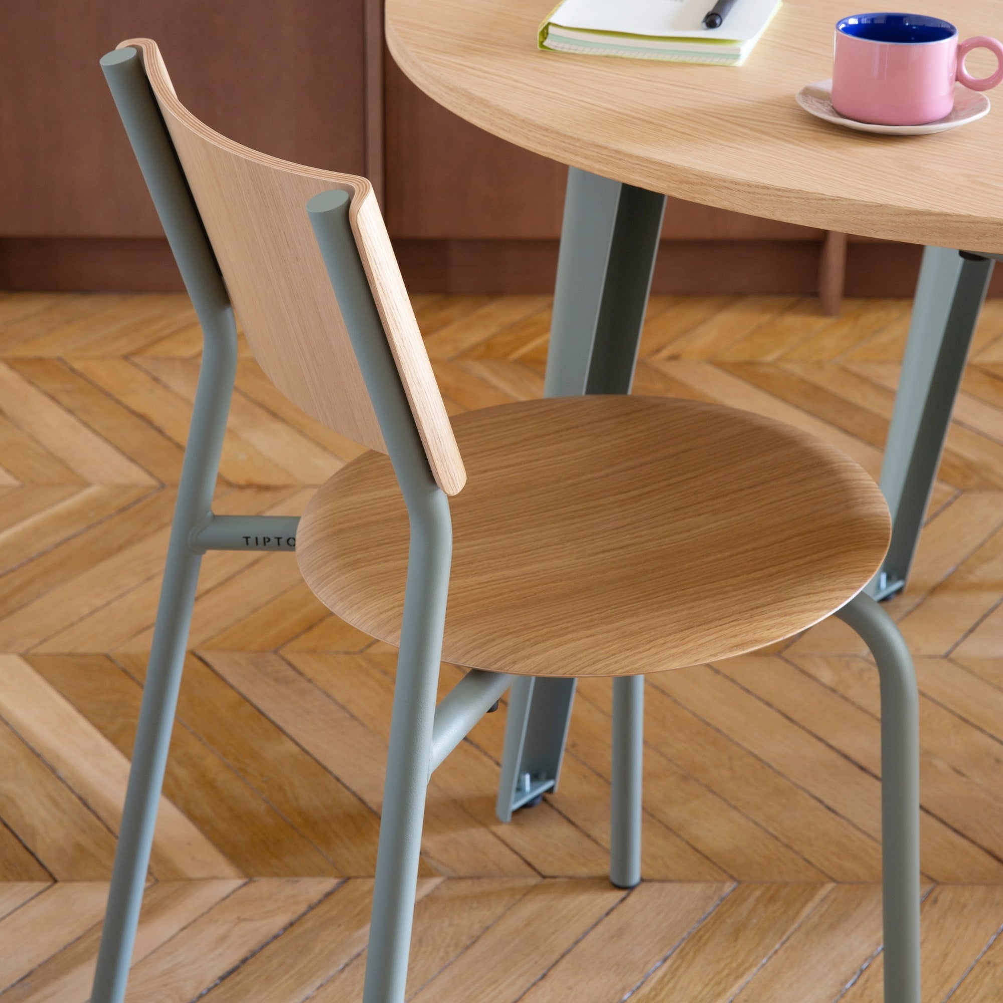 Chaise en bois et acier, modèle SSD de la marque Tiptoe, couleur gris, placée autour d’une table en bois, avec une tasse rose et un carnet posés dessus, sur un sol en parquet.