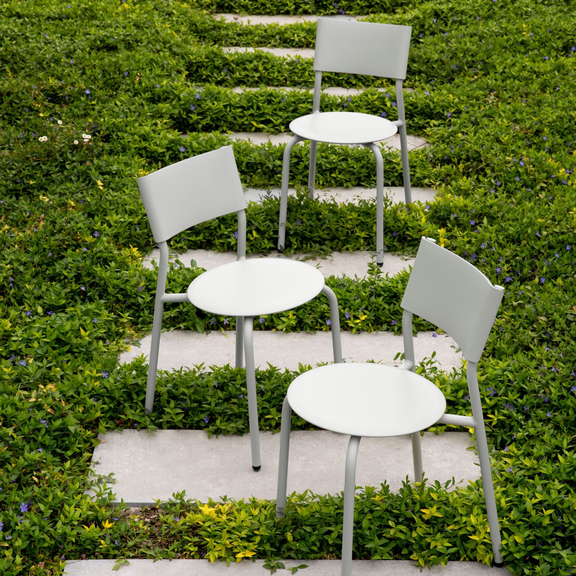 Trois chaises d’extérieur en métal et plastique recyclé gris clair, modèle SSDr, disposées sur un chemin en pierre entouré de verdure.