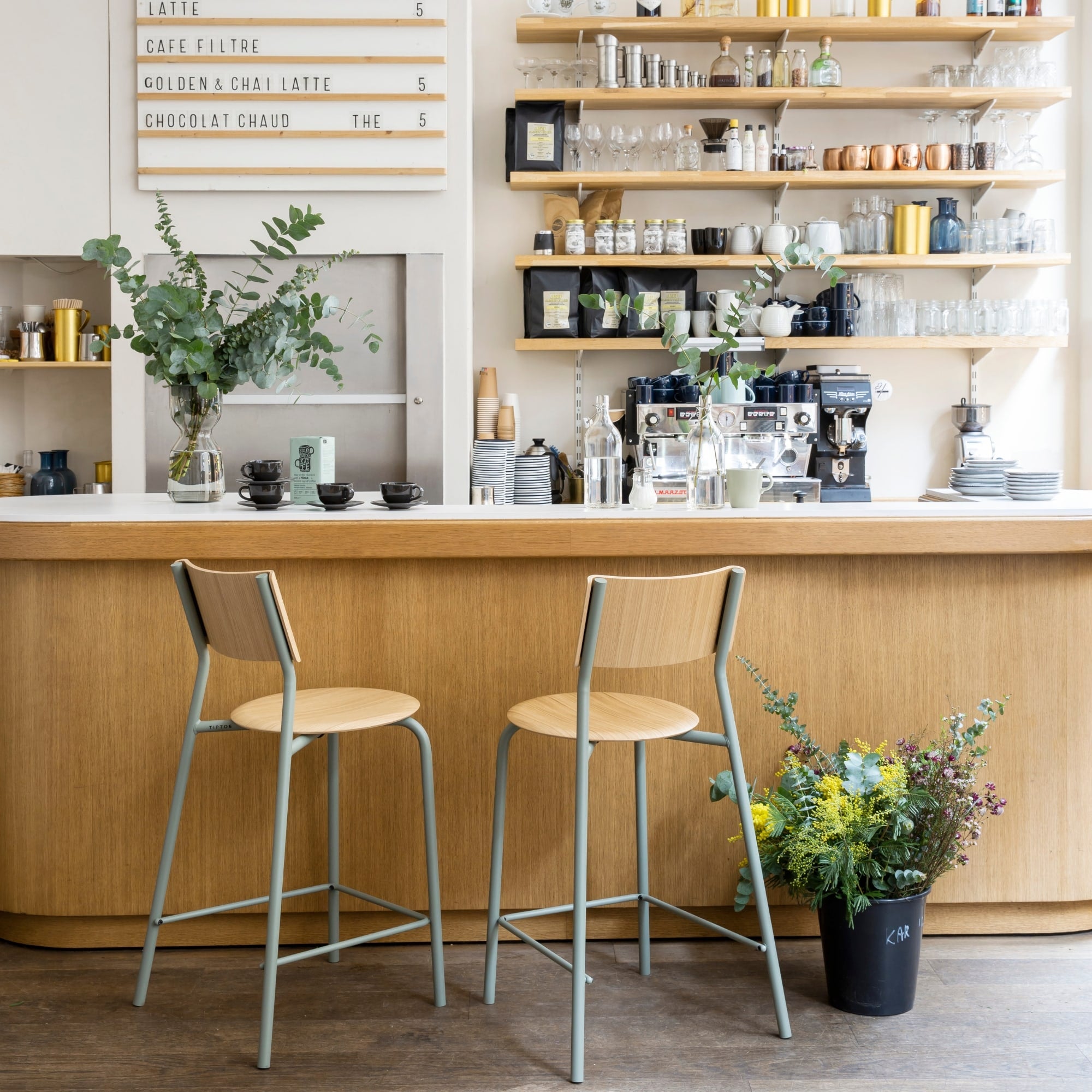 Deux chaises hautes en bois clair avec piètement métallique gris eucalyptus devant un comptoir en bois dans un café lumineux.