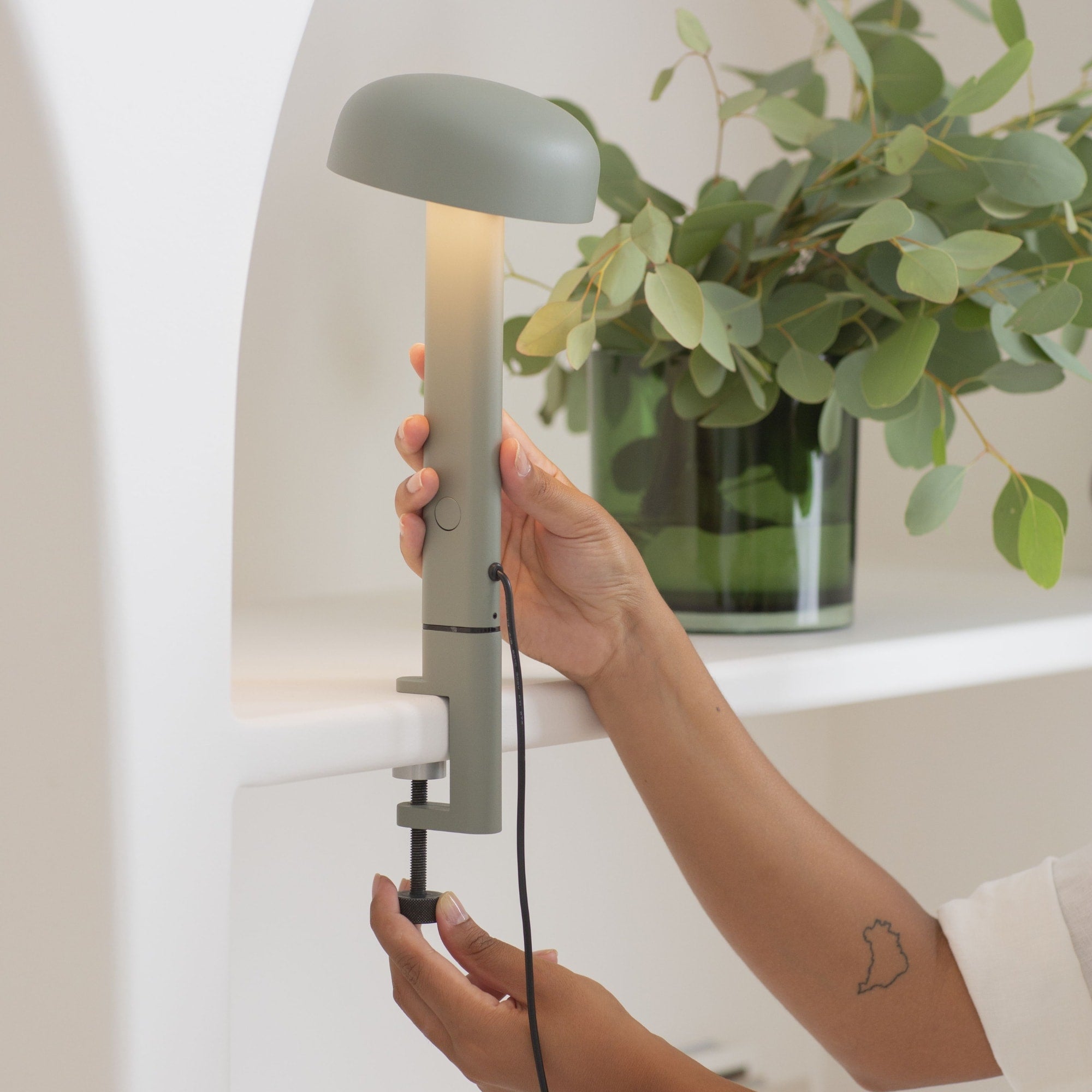Lampe à pince gris eucalyptus, modèle Nod de Tiptoe, vue en gros plan montrant une main ajustant la lampe. Le bras est fixé sur une étagère, avec l’abat-jour circulaire allumé, orienté vers le bas, et la lumière non visible.