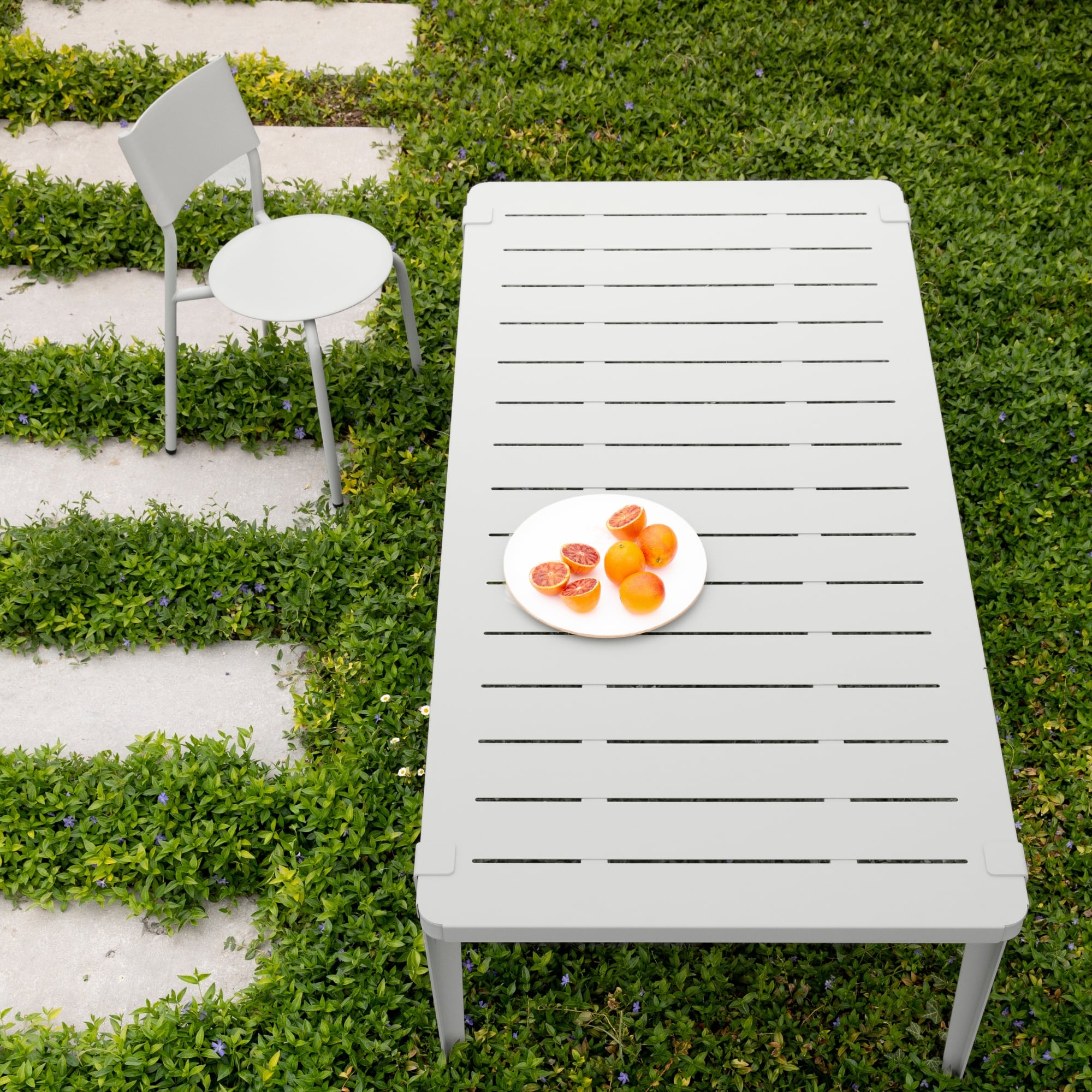 Table de jardin grise Tiptoe en extérieur, vue du dessus, avec une assiette de fruits posée dessus et une chaise assortie sur une pelouse en damier minéral et végétal.
