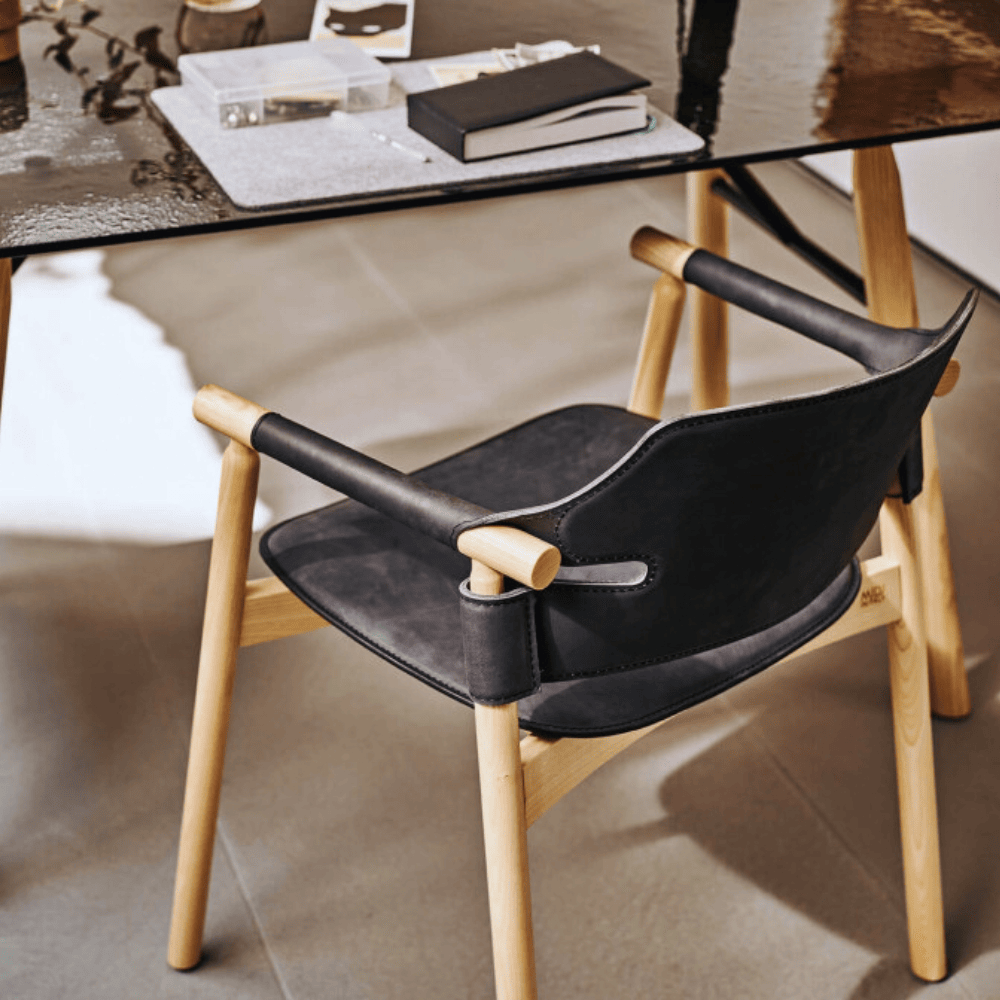 Fauteuil en bois clair avec assise et dossier en cuir noir, placé devant un bureau au plateau en verre.