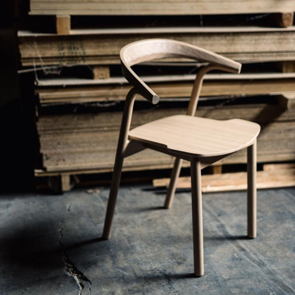 Chaise en bois clair, posée sur un sol en béton brut, devant un stock de planches en bois.