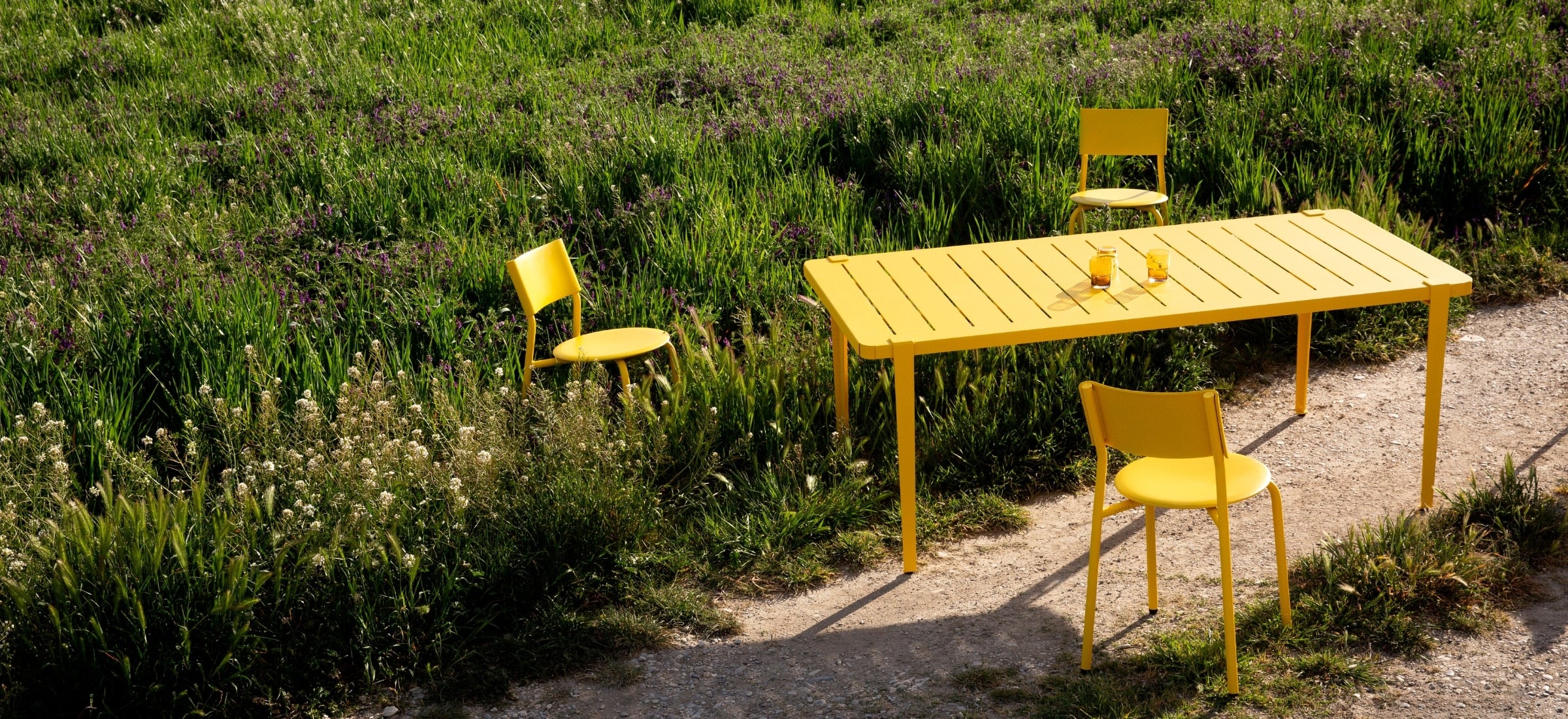 Table et chaises d’extérieur en métal et plastique recyclé jaune Tiptoe, installées sur un sentier entouré de verdure et de fleurs sauvages.