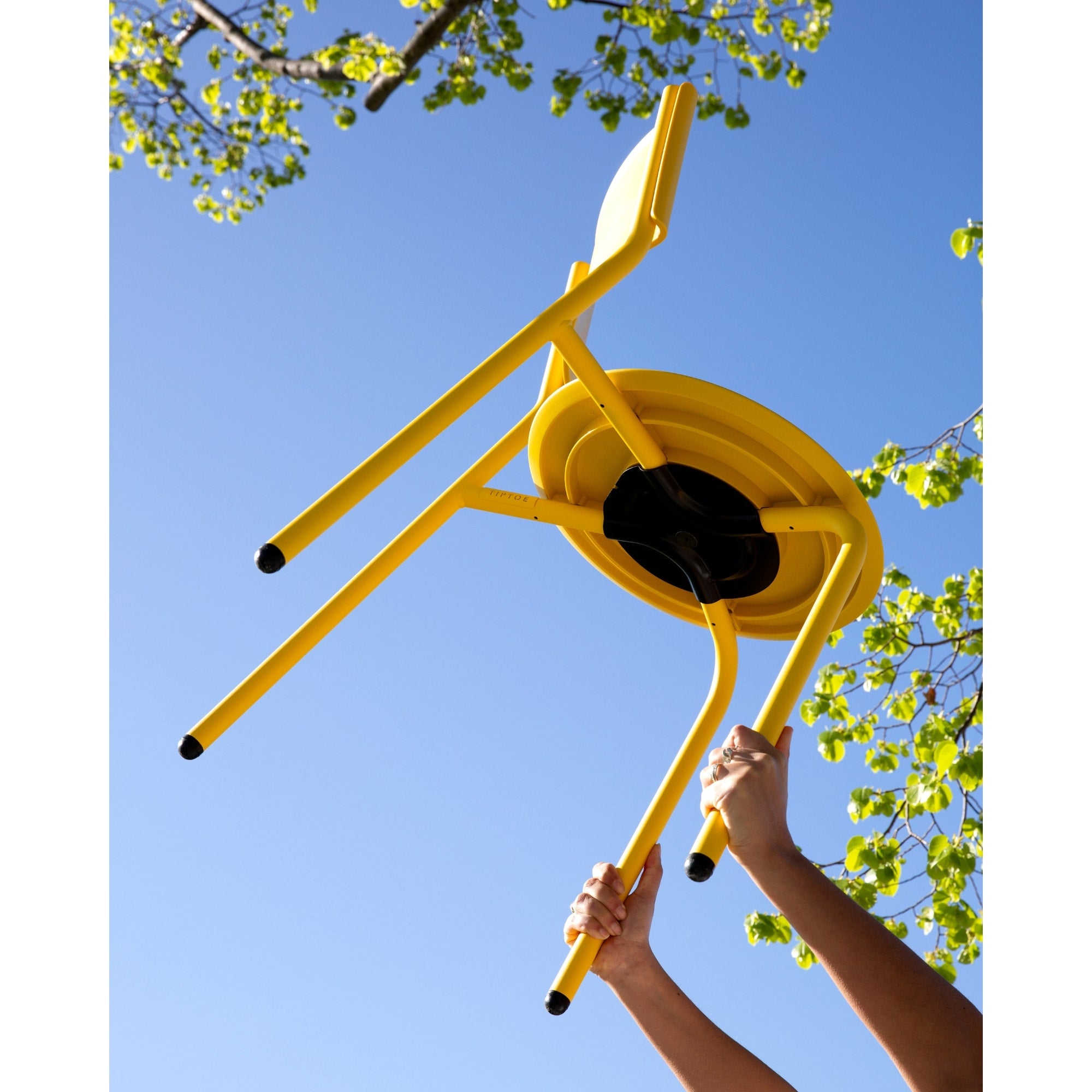 Chaise en métal et plastique recyclé jaune Tiptoe, vue du dessous, tenue à bout de bras sous un ciel bleu.