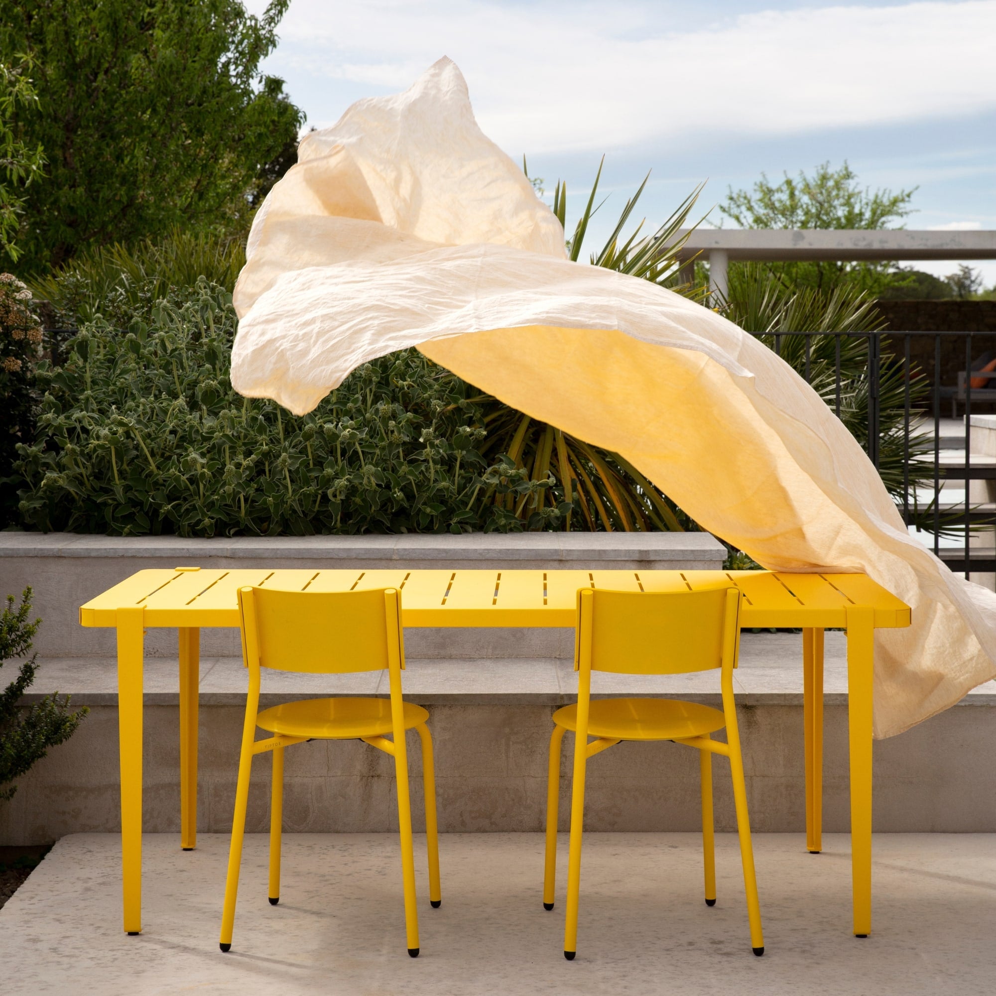 Table de jardin jaune Tiptoe accompagnée de deux chaises assorties, sur une terrasse végétalisée avec un voile flottant au vent.