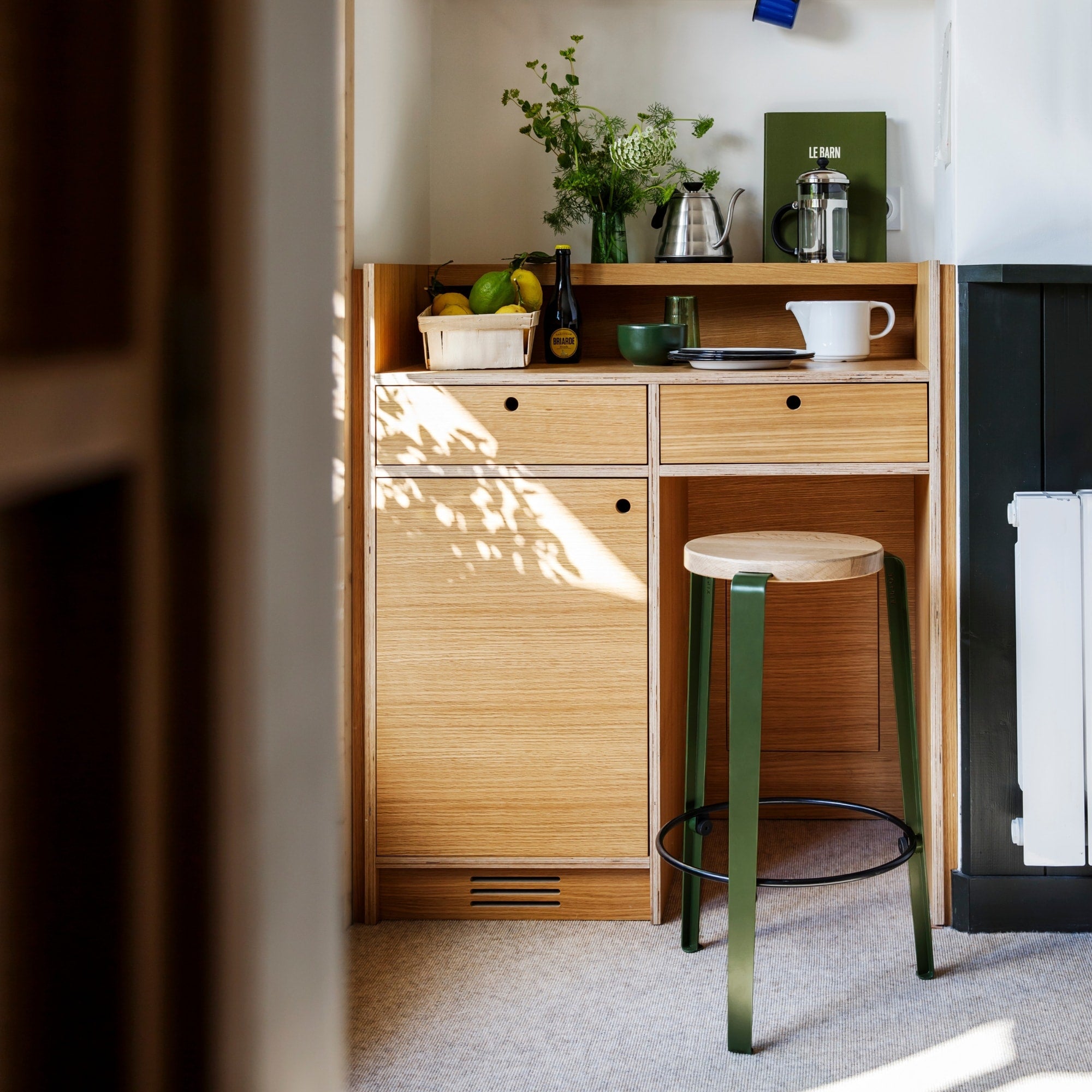 Tabouret de bar Lou avec assise en bois clair et pieds en acier vert, placé devant un meuble de cuisine en bois naturel.