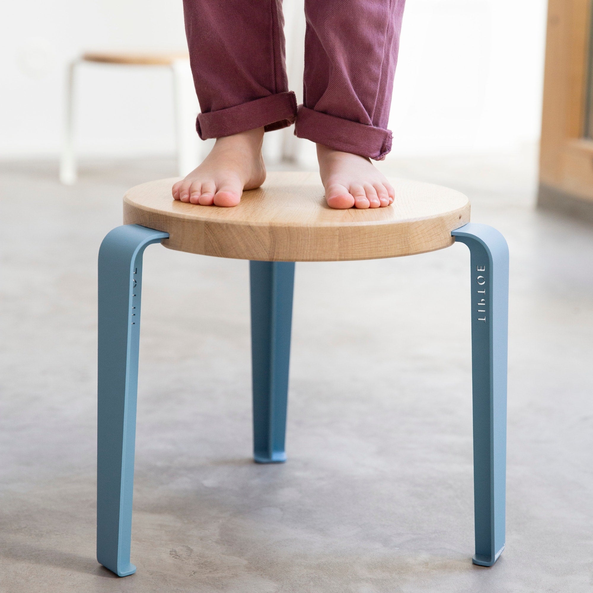 Tabouret enfant Tiptoe en bois et acier bleu baleine, sur lequel un enfant se tient debout pieds nus, sol en béton