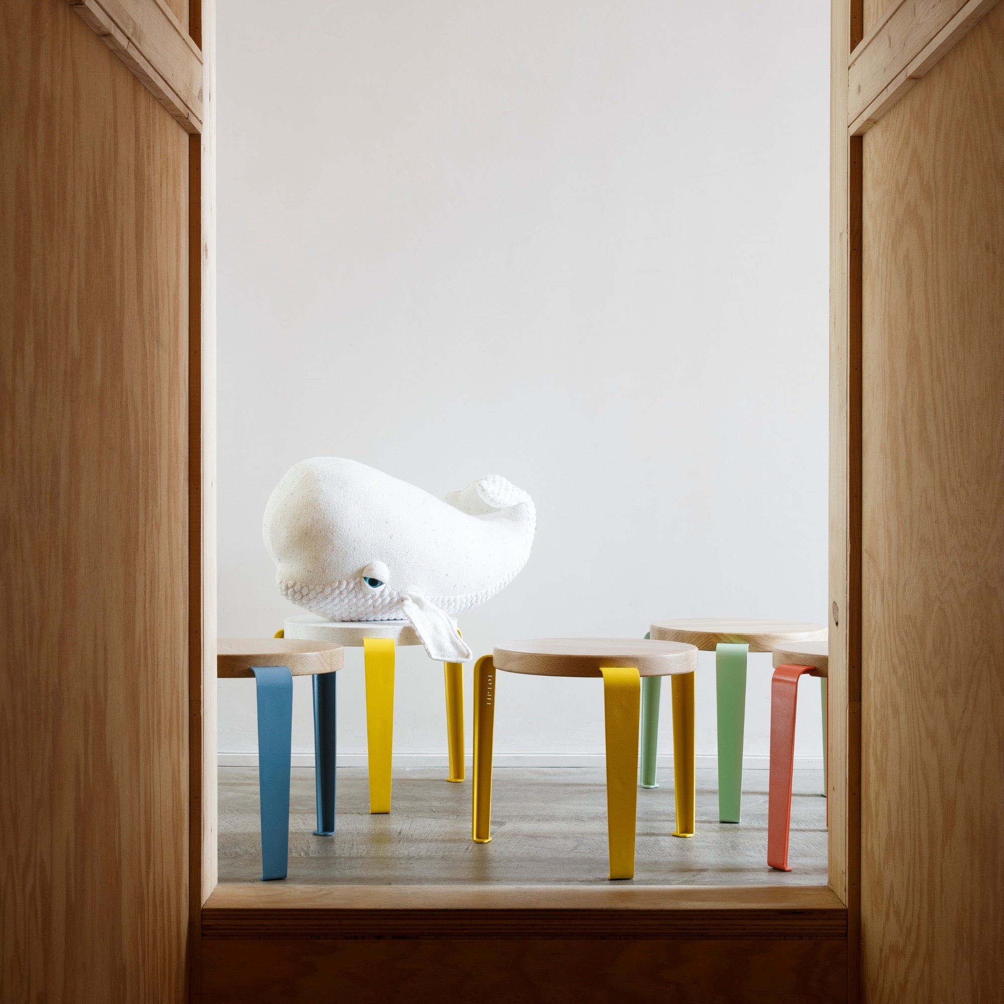 Tabourets enfant Tiptoe en bois et acier coloré, alignés sous une peluche blanche en forme de baleine, sol en bois clair