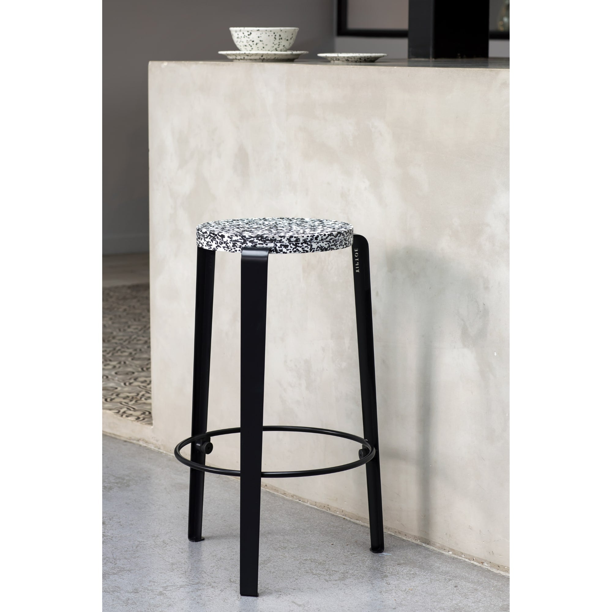Tabouret de comptoir Lou avec assise en plastique recyclé noir et blanc, pieds noirs, devant un bar en béton.