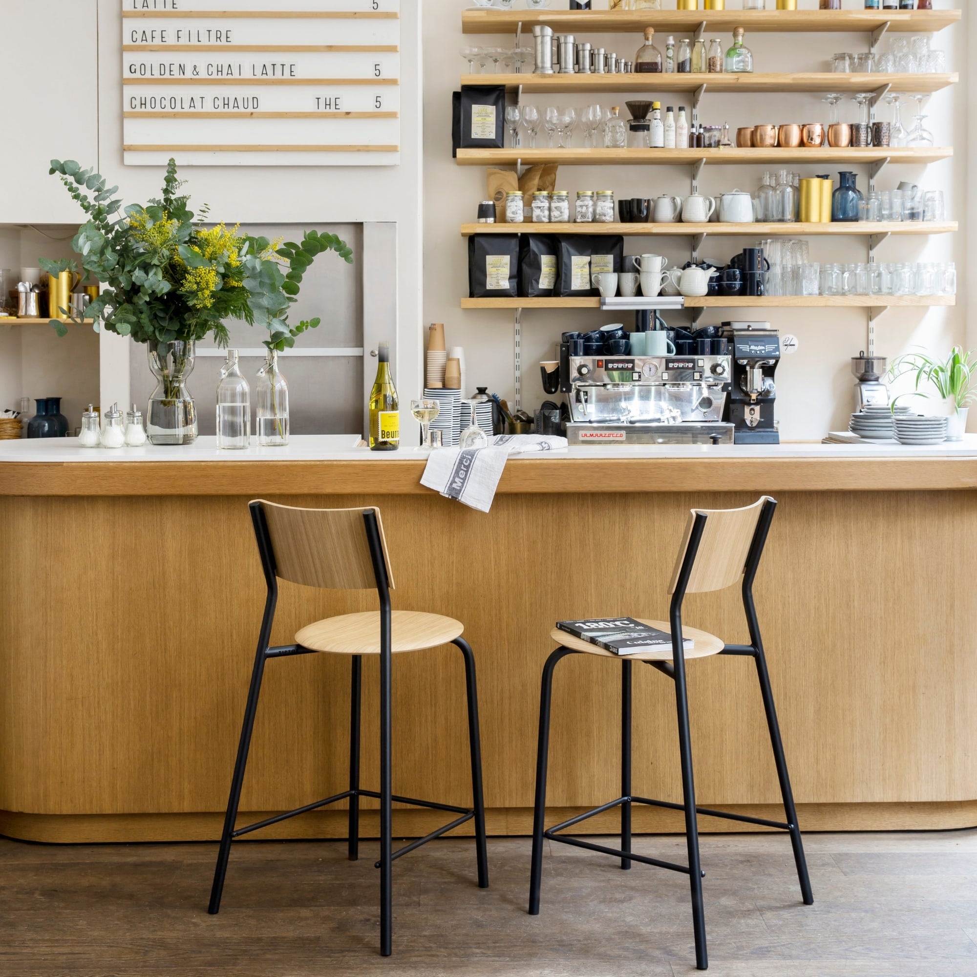 Deux chaises hautes en bois clair avec piètement métallique noir devant un comptoir en bois dans un café lumineux avec étagères décoratives.