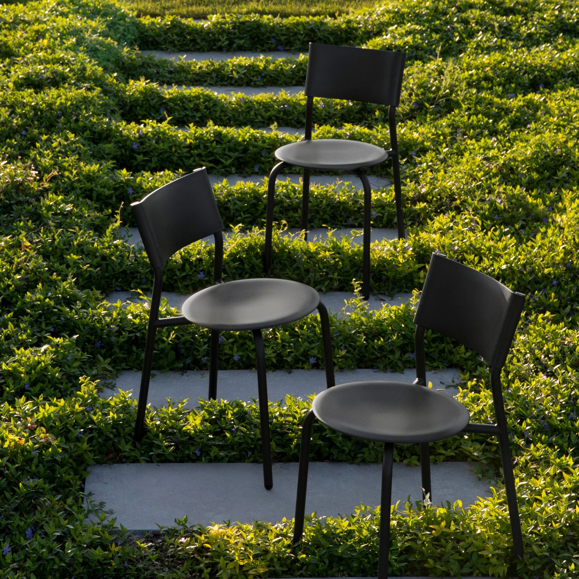 Trois chaises de jardin en métal et plastique recyclé noir disposées sur un chemin extérieur verdoyant.