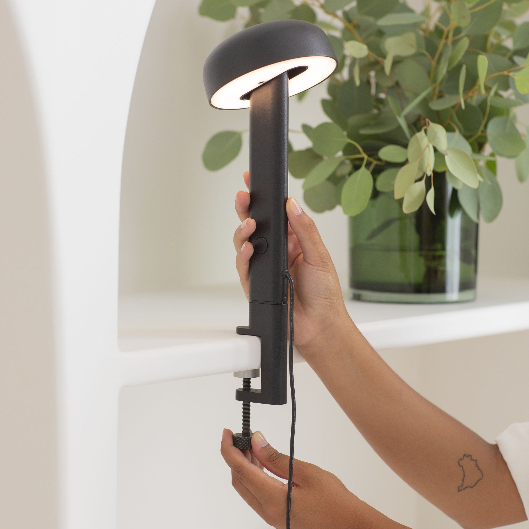 Lampe à pince noire, modèle Nod de Tiptoe, vue en gros plan montrant une main ajustant la lampe. Le bras est fixé sur une étagère, avec l’abat-jour circulaire allumé, orienté vers le bas, et la lumière visible.