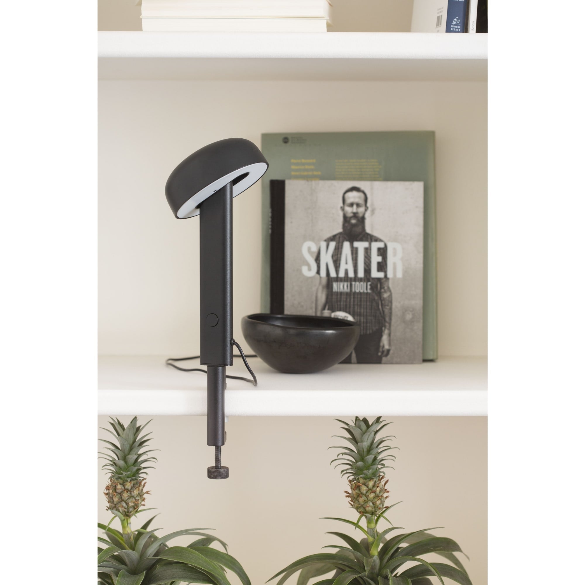 Lampe à pince noire, modèle Nod de Tiptoe, vue latérale. Le bras fin soutient un abat-jour circulaire, avec un livre et un bol noir en arrière-plan, et des plantes en pot sur l’étagère.