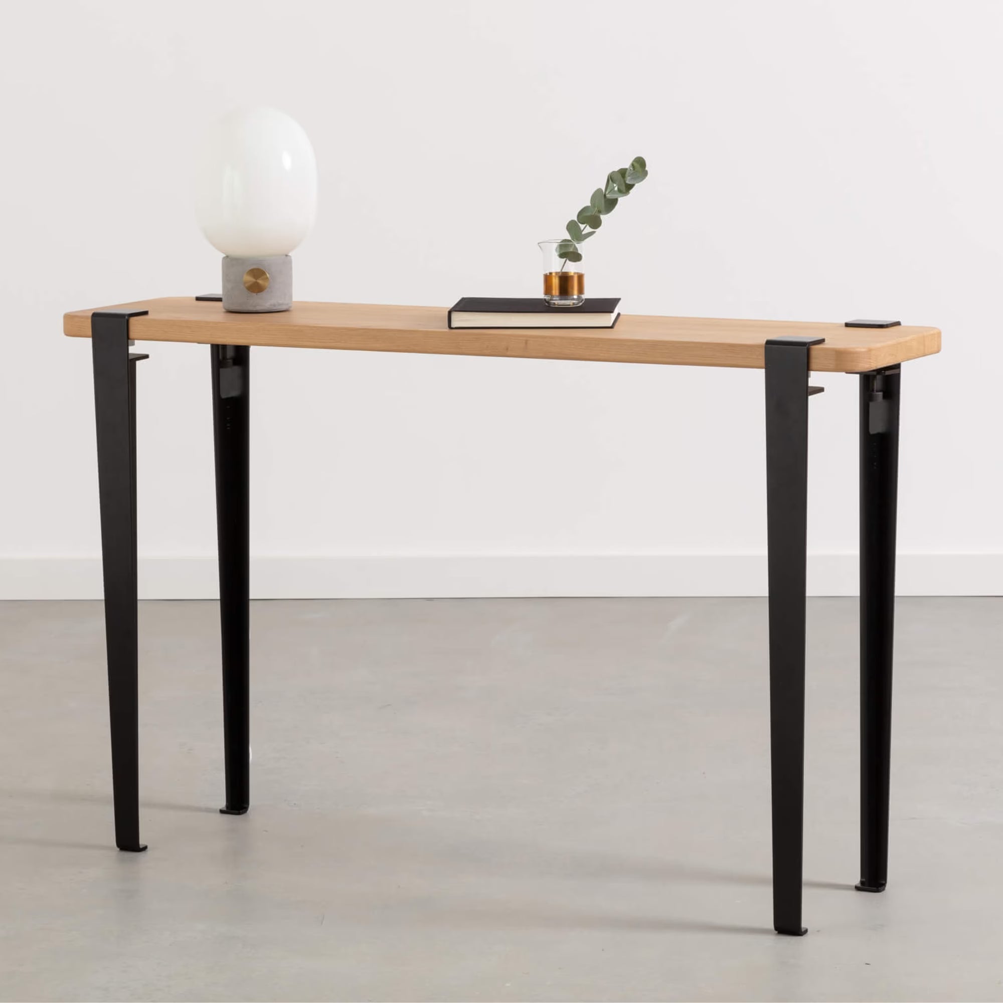 Petite console en bois et métal noir, plateau chêne clair, décorée d’une lampe et d’un carnet posé sur fond neutre.