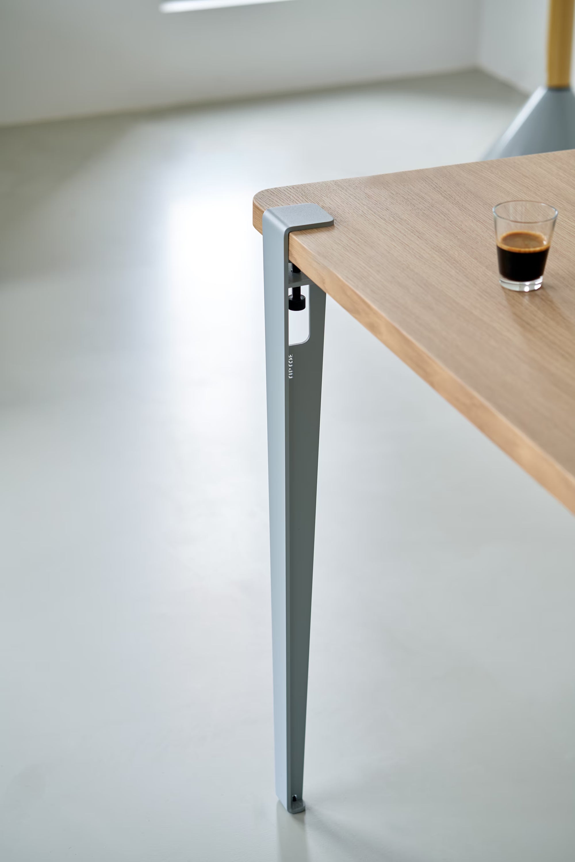 Pied de table en métal bleu pâle Tiptoe, fixé à un plateau en bois clair ; vis de serrage apparente et petit verre d’expresso posé.