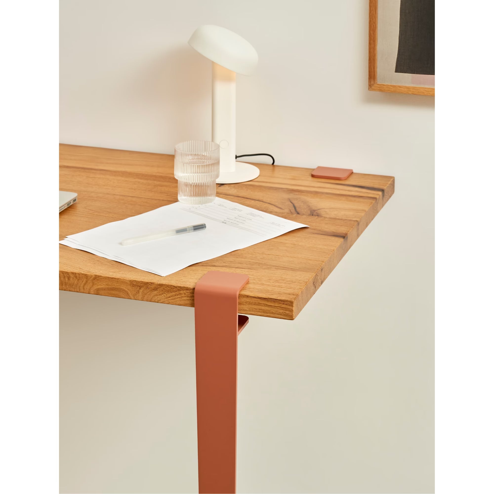Bureau en bois clair avec pied métallique brun cannelle, lampe blanche et feuille posées sur le plateau.
