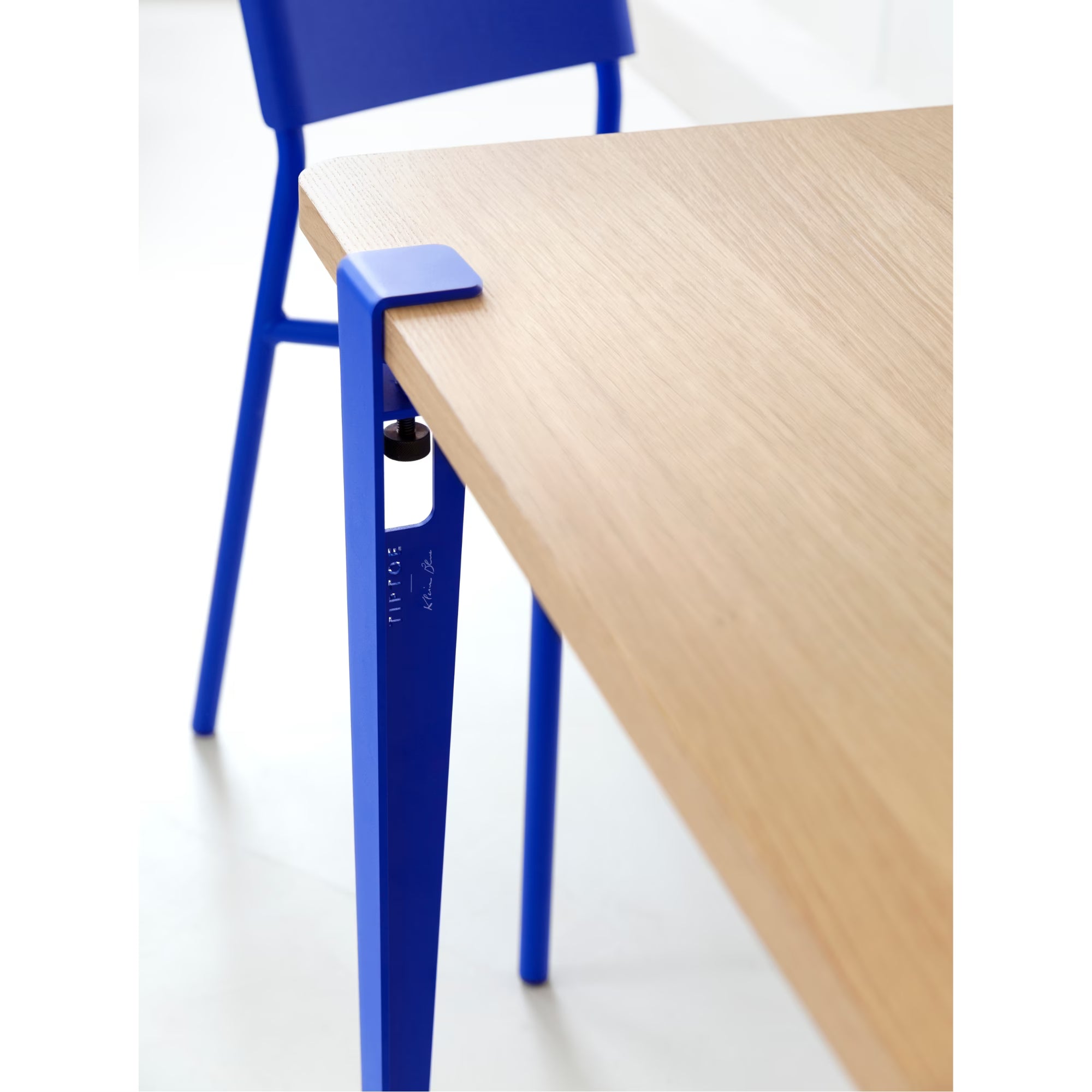 Vue rapprochée d’un pied de table bleu Klein fixé à un plateau en bois clair, chaise assortie en arrière-plan.