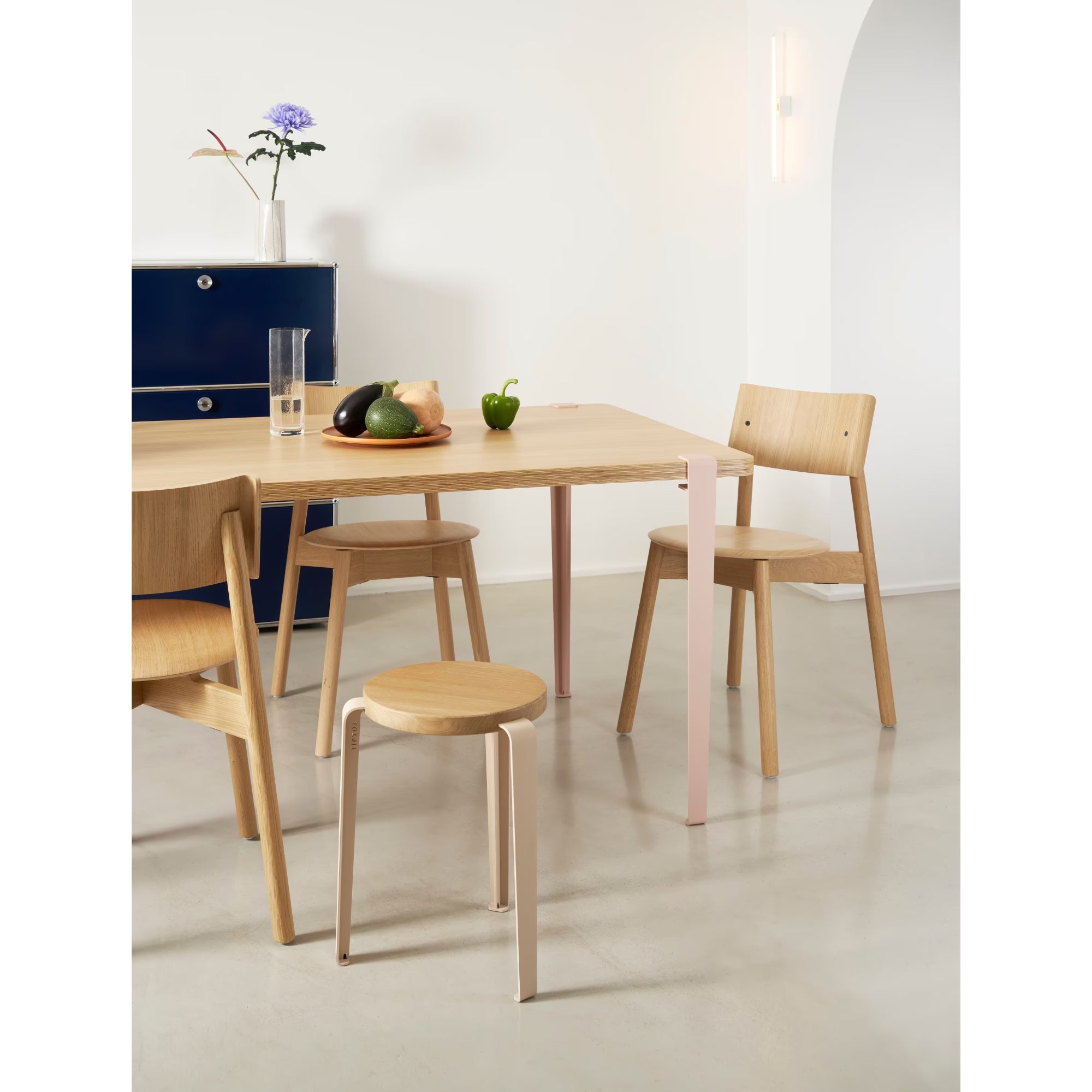 Salle à manger lumineuse avec table en bois clair, chaises et tabouret assortis, buffet bleu au fond et détail rose pâle discret.