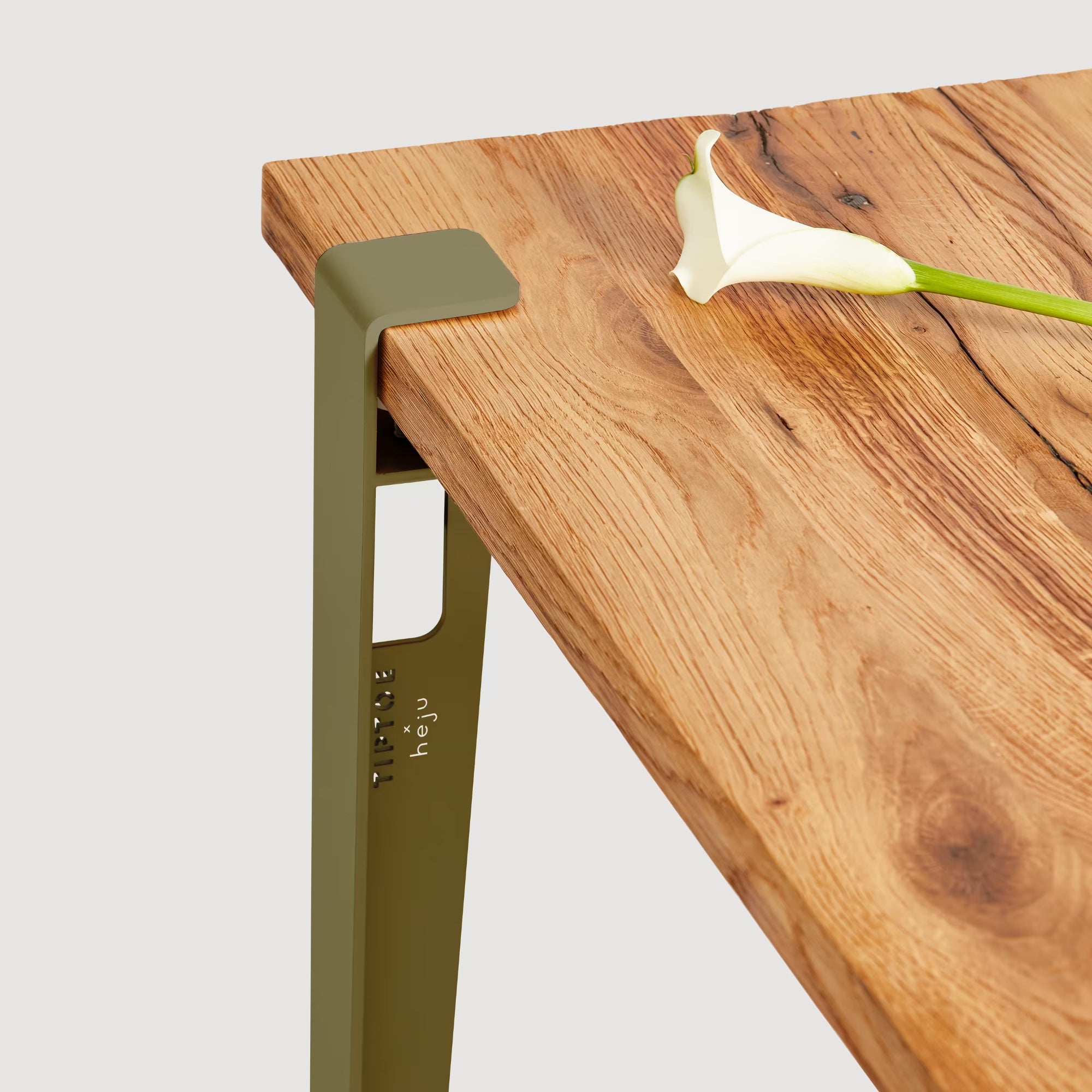 Vue rapprochée d’un pied de table vert bruyère fixé à un plateau en bois, avec une fleur blanche posée dessus.