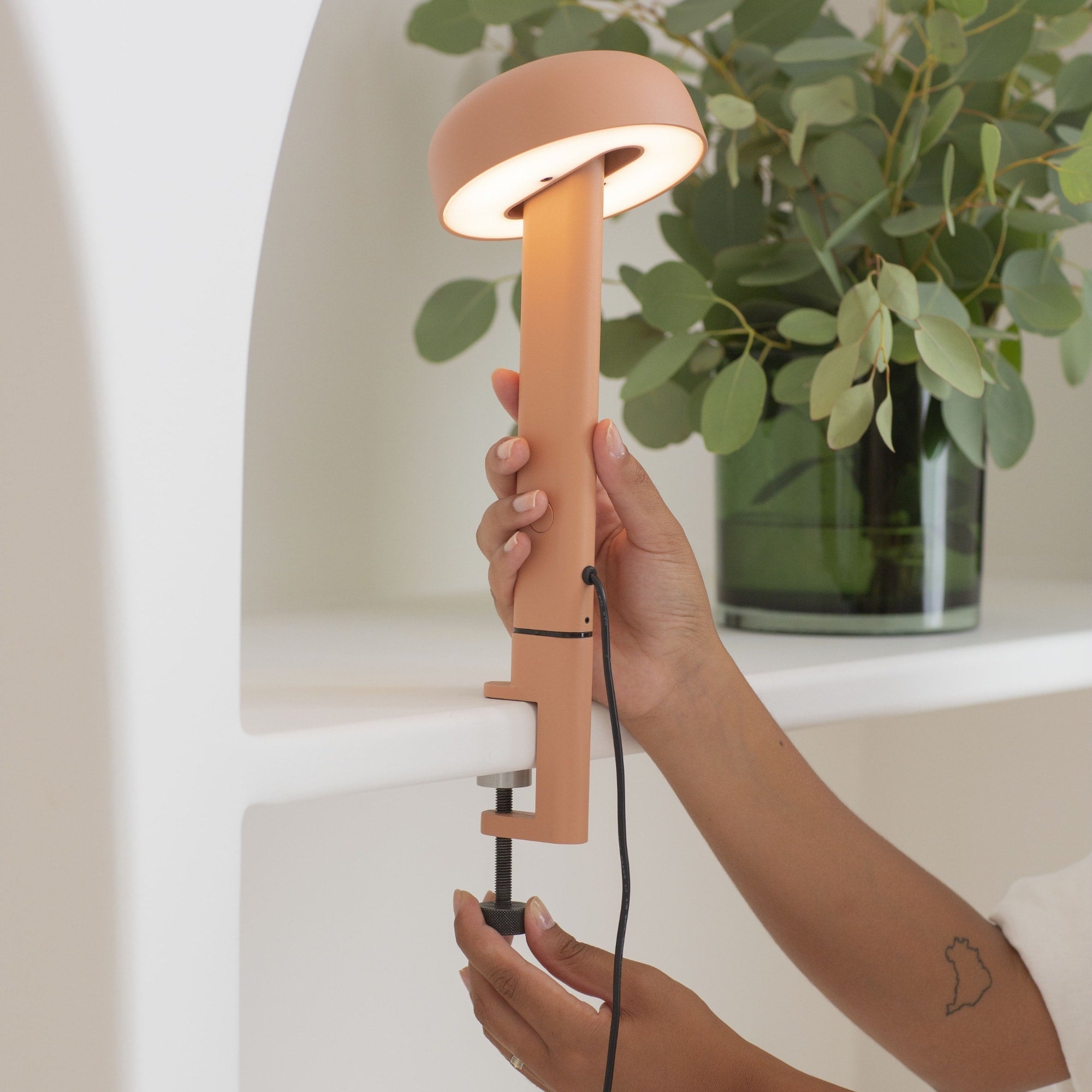 Lampe à pince rose, modèle Nod de Tiptoe, vue en gros plan montrant une main ajustant la lampe. Le bras est fixé sur une étagère, avec l’abat-jour circulaire allumé, orienté vers le bas, et la lumière visible.