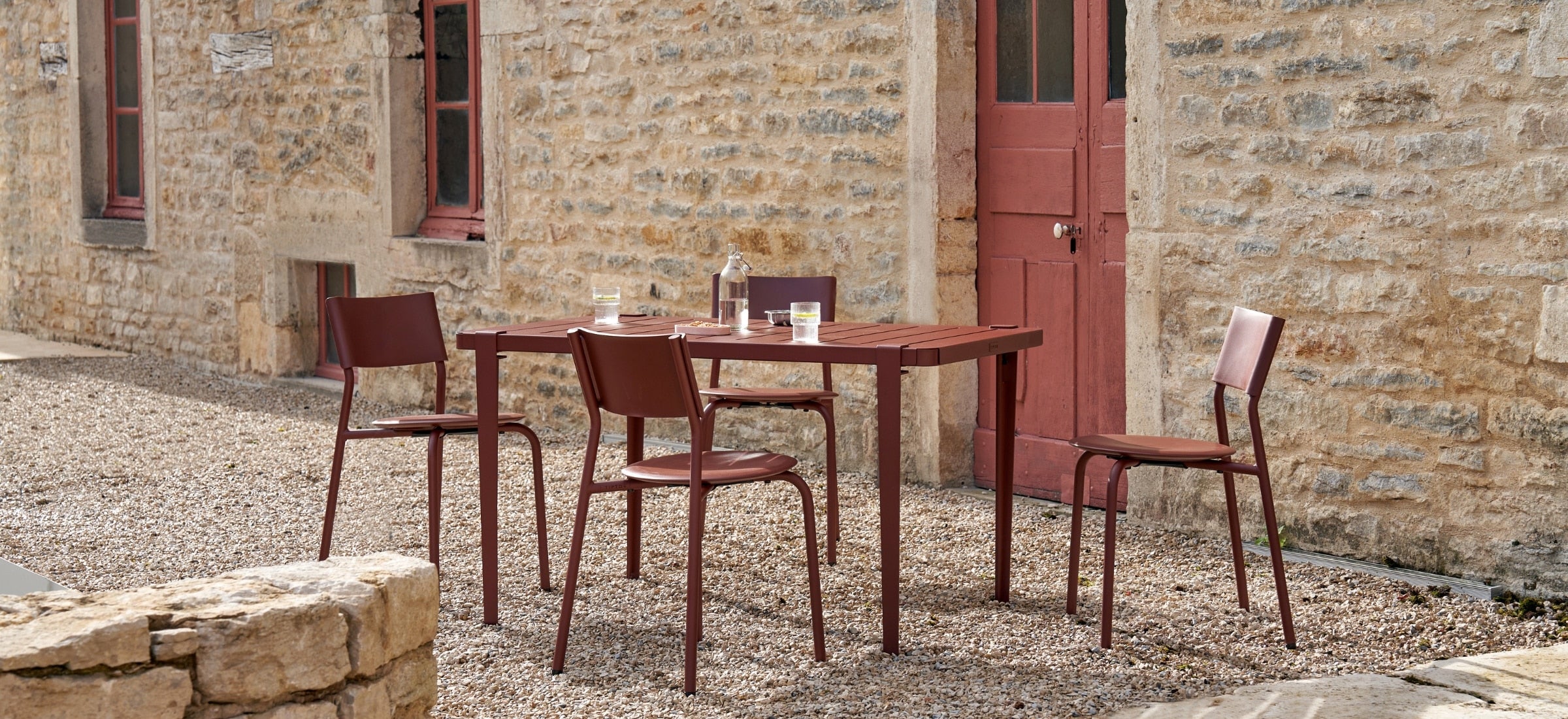 Ensemble de table et chaises en métal et plastique recyclé rouge brique Tiptoe, installé sur une terrasse en pierre devant une façade en pierre naturelle.