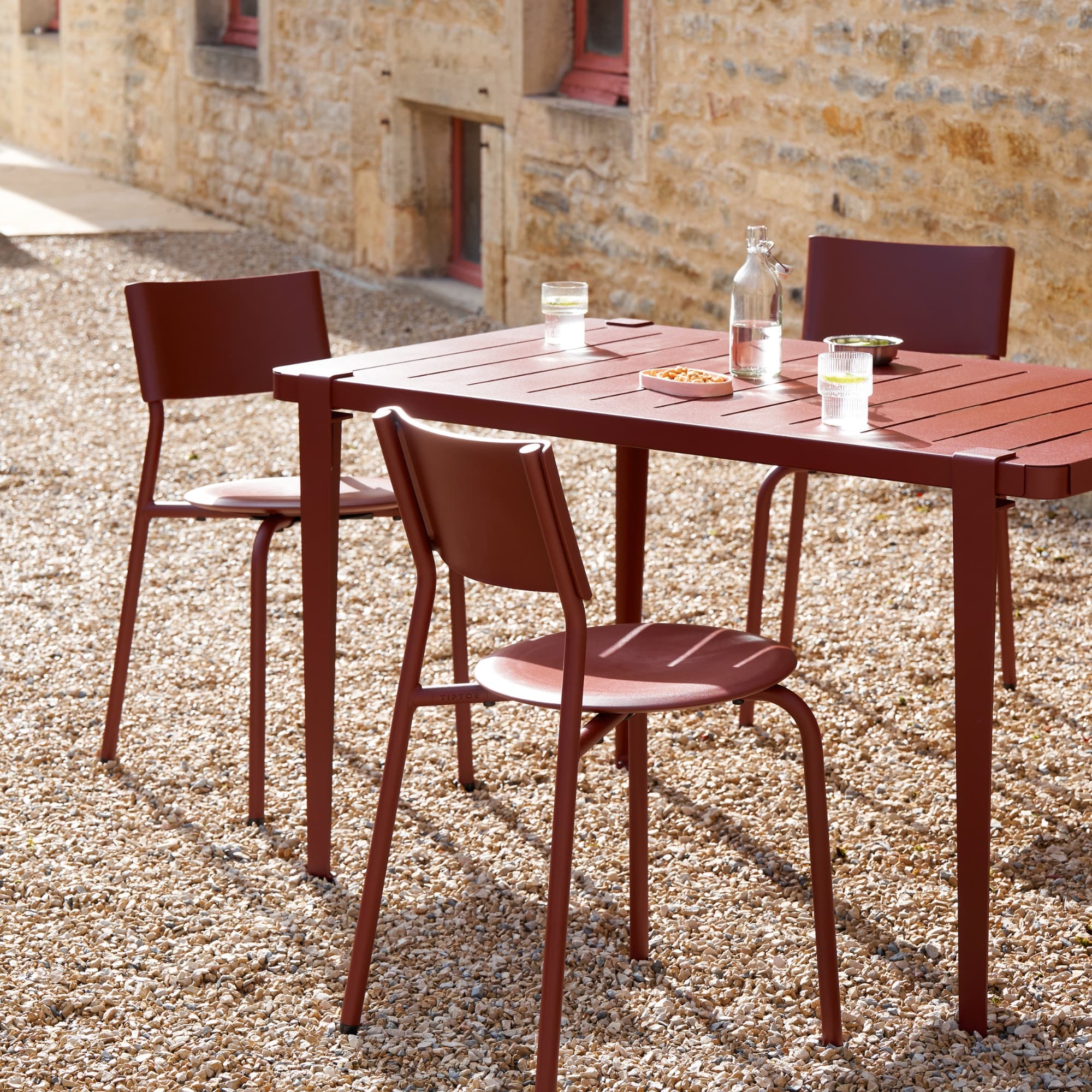 Chaises de jardin en métal et plastique recyclé rouge autour d’une table assortie, sur une cour en gravier et maison en pierre.