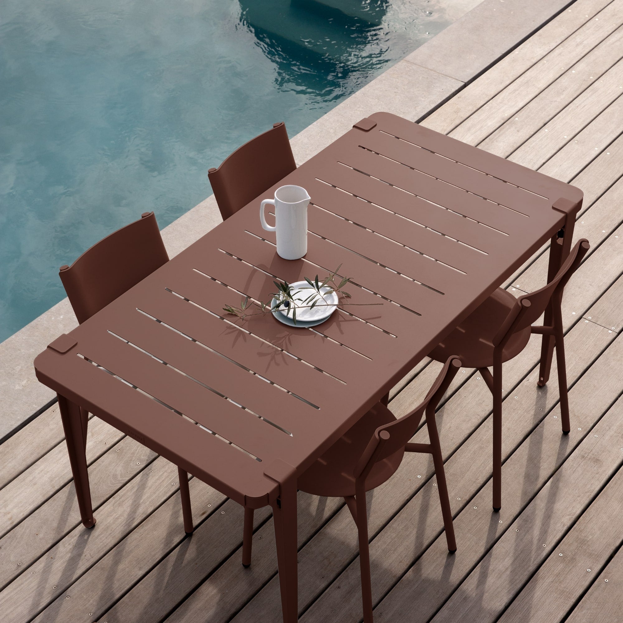 Table de jardin rouge Tiptoe avec six chaises assorties, installée sur une terrasse en bois au bord d’une piscine, vue en plongée.