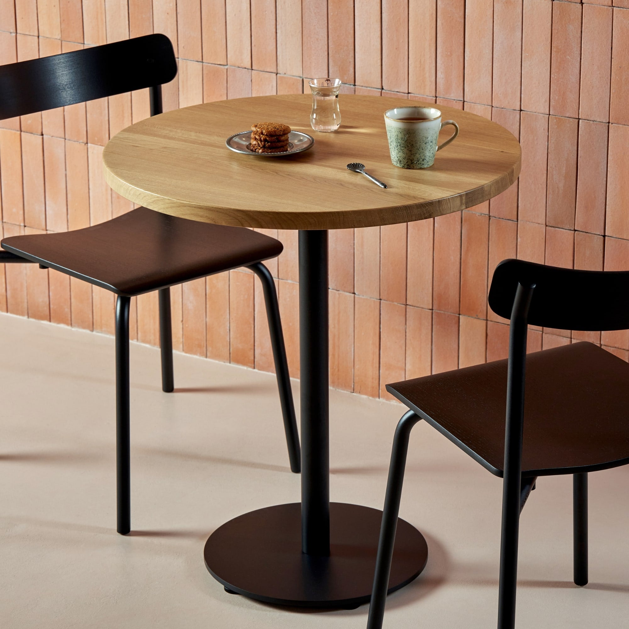 Table bistrot ronde en bois avec pied central noir, entourée de deux chaises noires dans un intérieur en briques rosées.